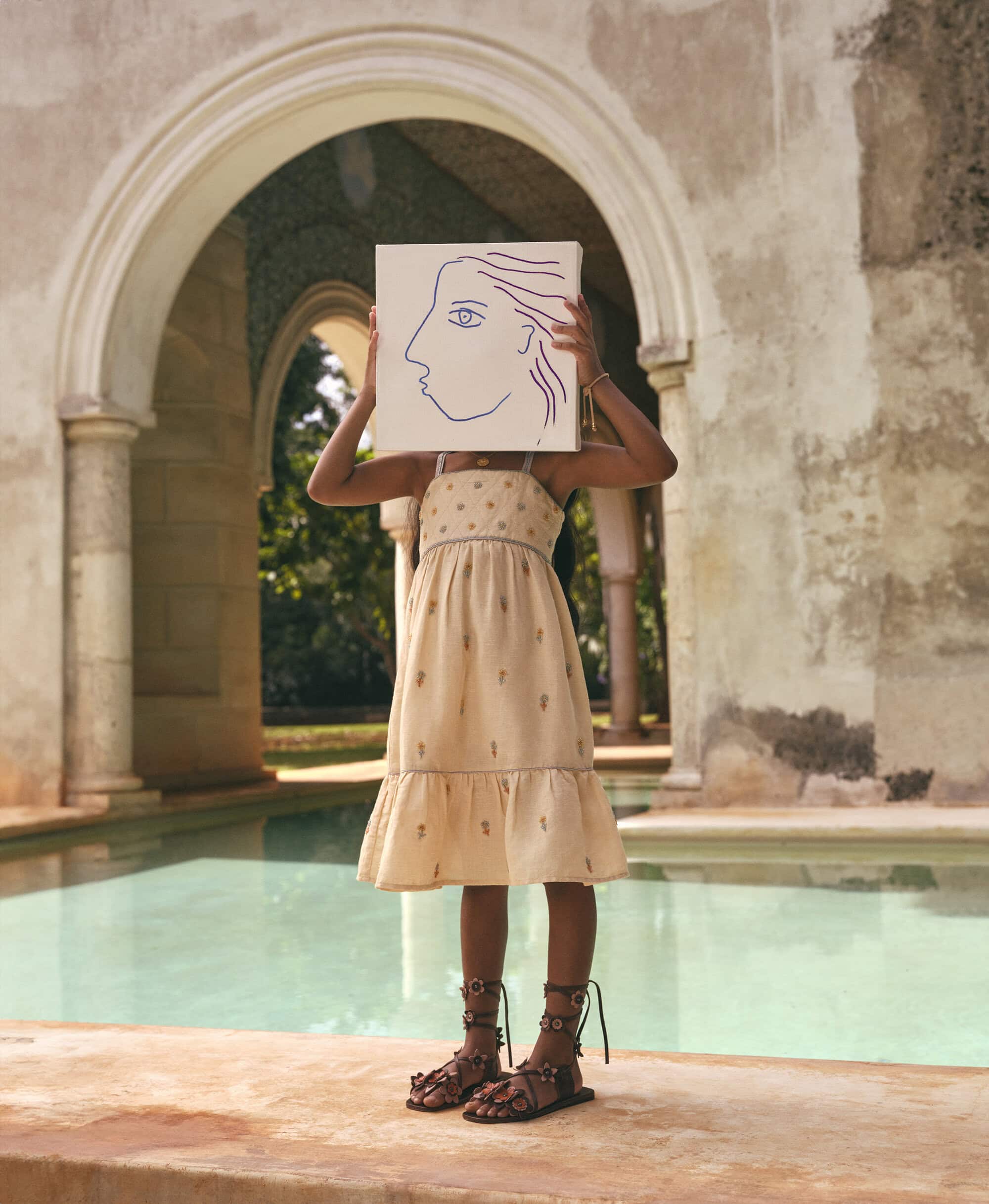 Young person in a dress stands by a pool, holding a drawing of a face in front of their face, with arched architecture in the background.