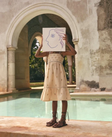Young person in a dress stands by a pool, holding a drawing of a face in front of their face, with arched architecture in the background.