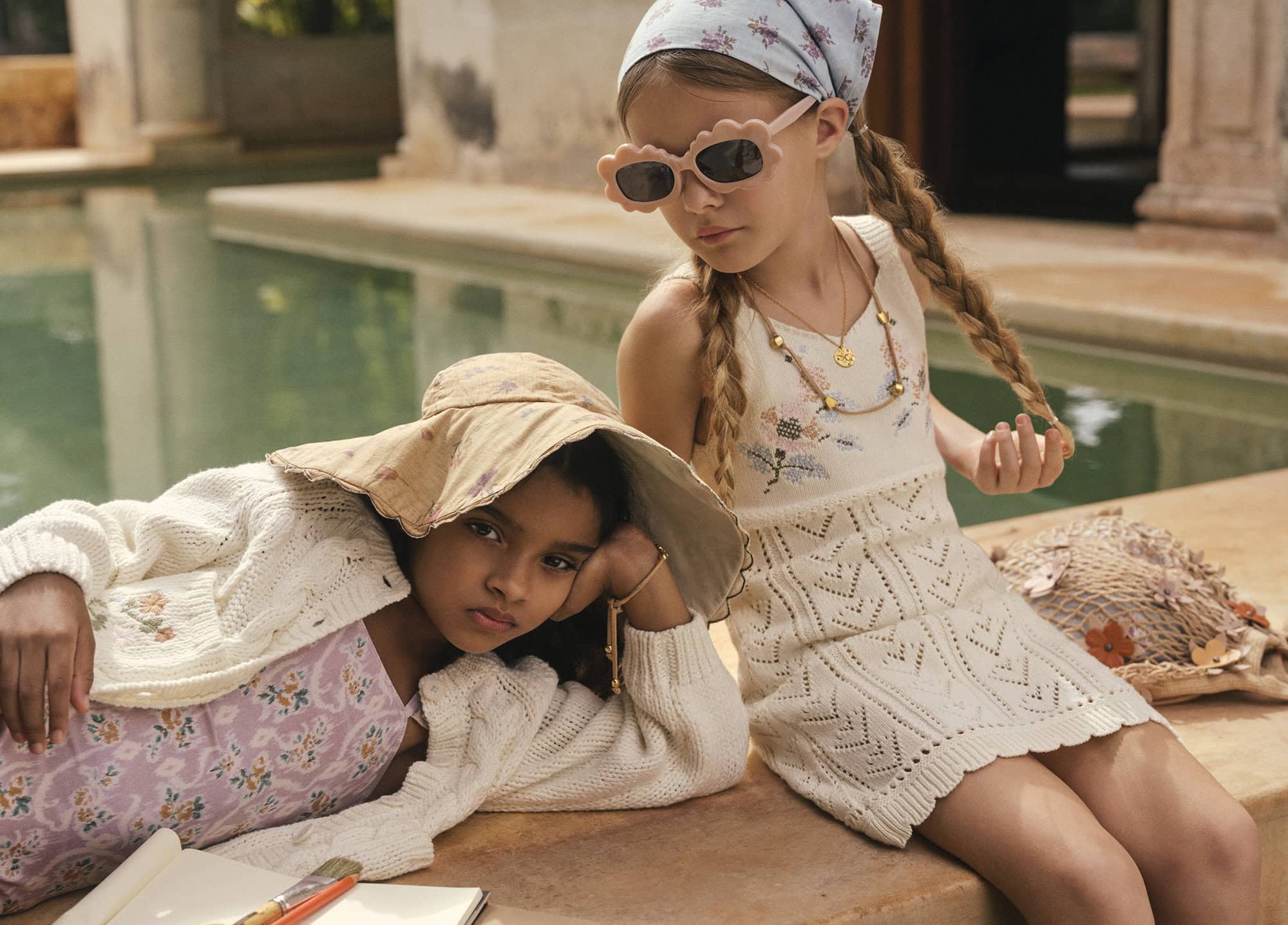 Two young girls dressed in vintage summer clothes sitting by a pool; one wears a floral headscarf and the other a large hat.