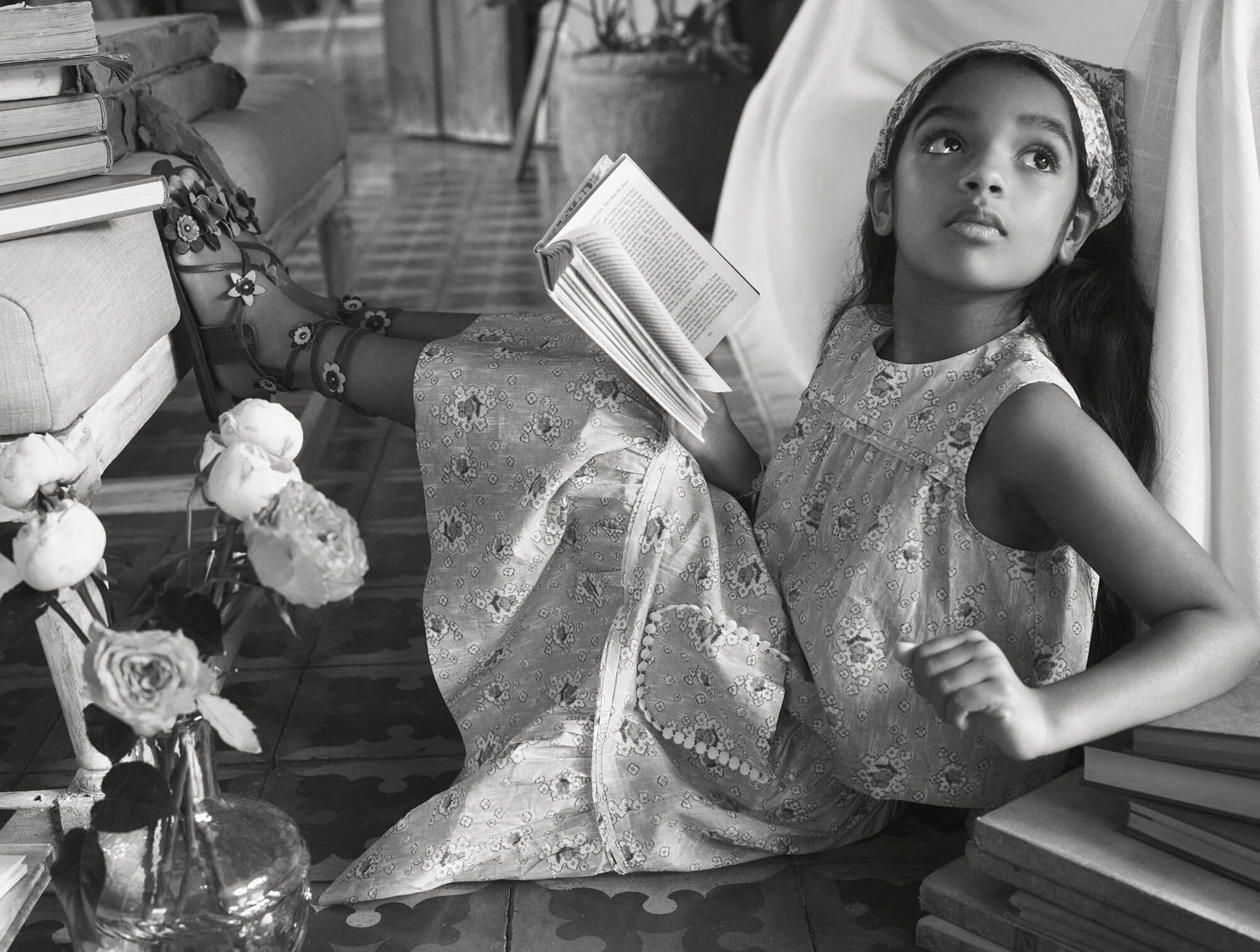 A young girl in a floral dress sits surrounded by books indoors, reading in a black and white photograph, wearing floral sandals.