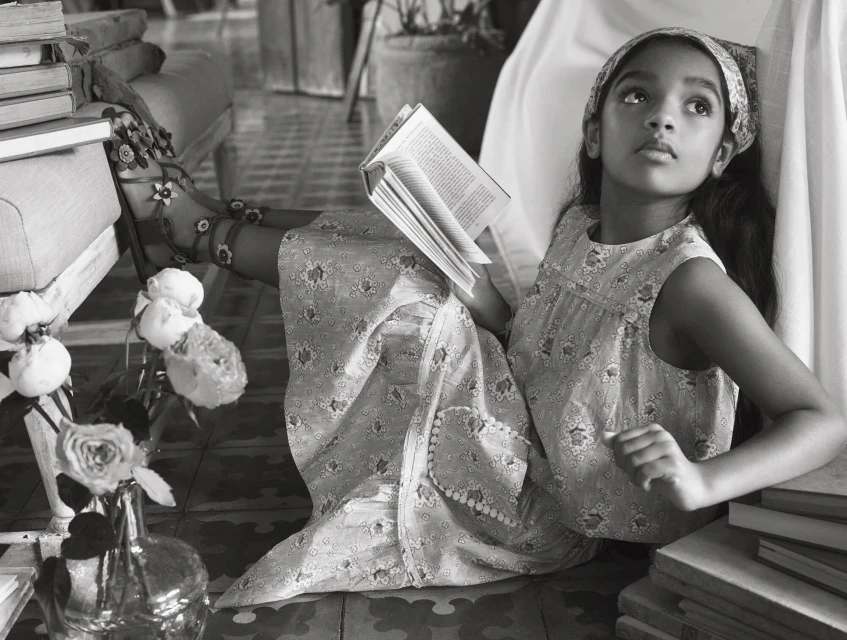 A young girl in a floral dress sits surrounded by books indoors, reading in a black and white photograph, wearing floral sandals.