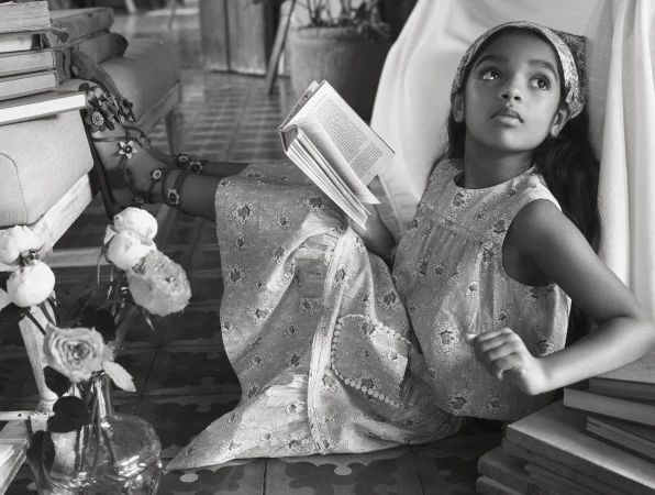 A young girl in a floral dress sits surrounded by books indoors, reading in a black and white photograph, wearing floral sandals.