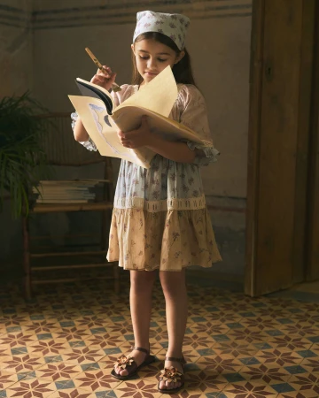 A young girl holding an open book and paintbrush, wearing a patterned dress and matching headscarf indoors.