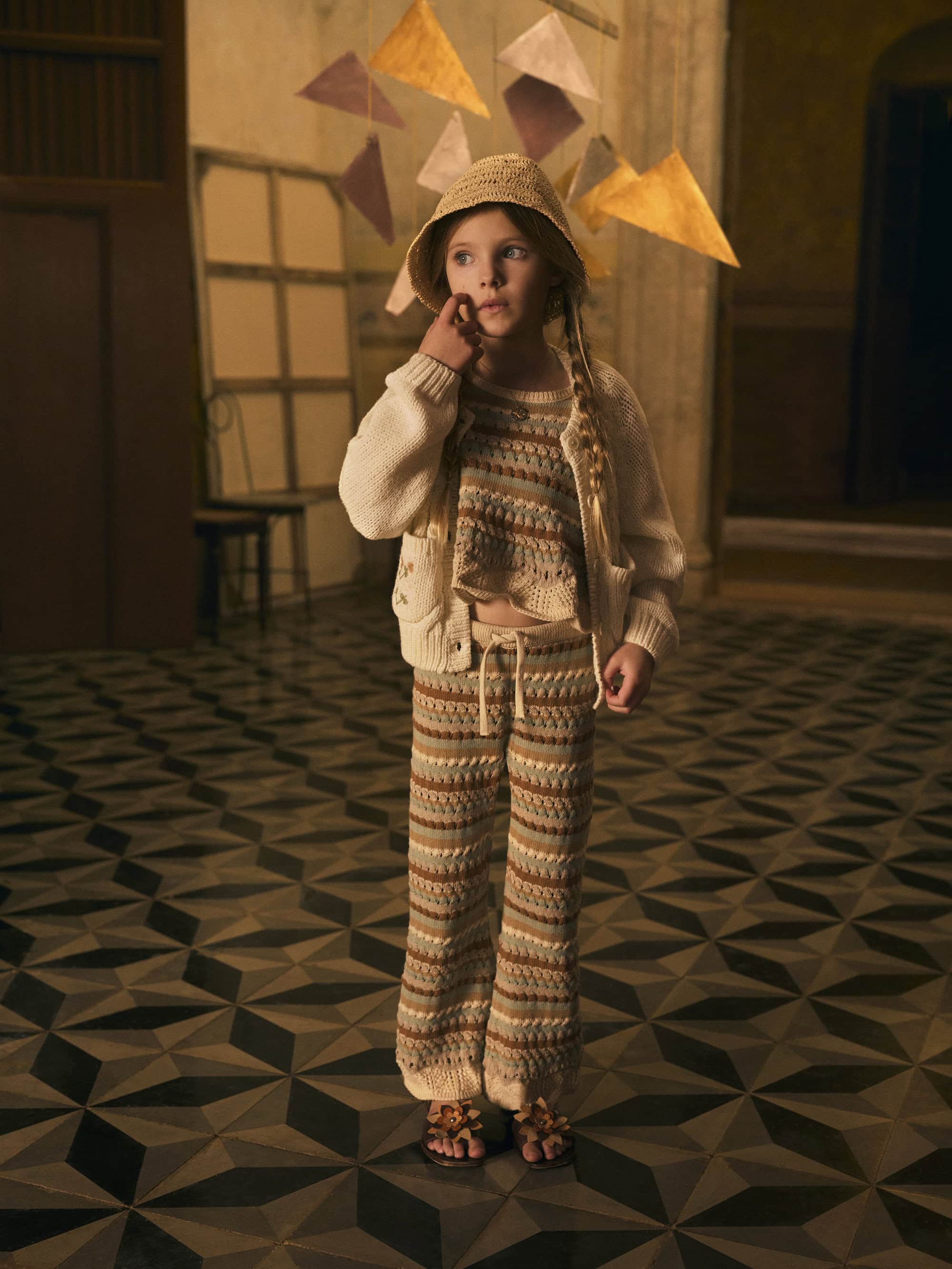 Young girl poses indoors wearing a knitted hat, cardigan, and pants. Decorative geometric shapes hang overhead, creating a whimsical effect.