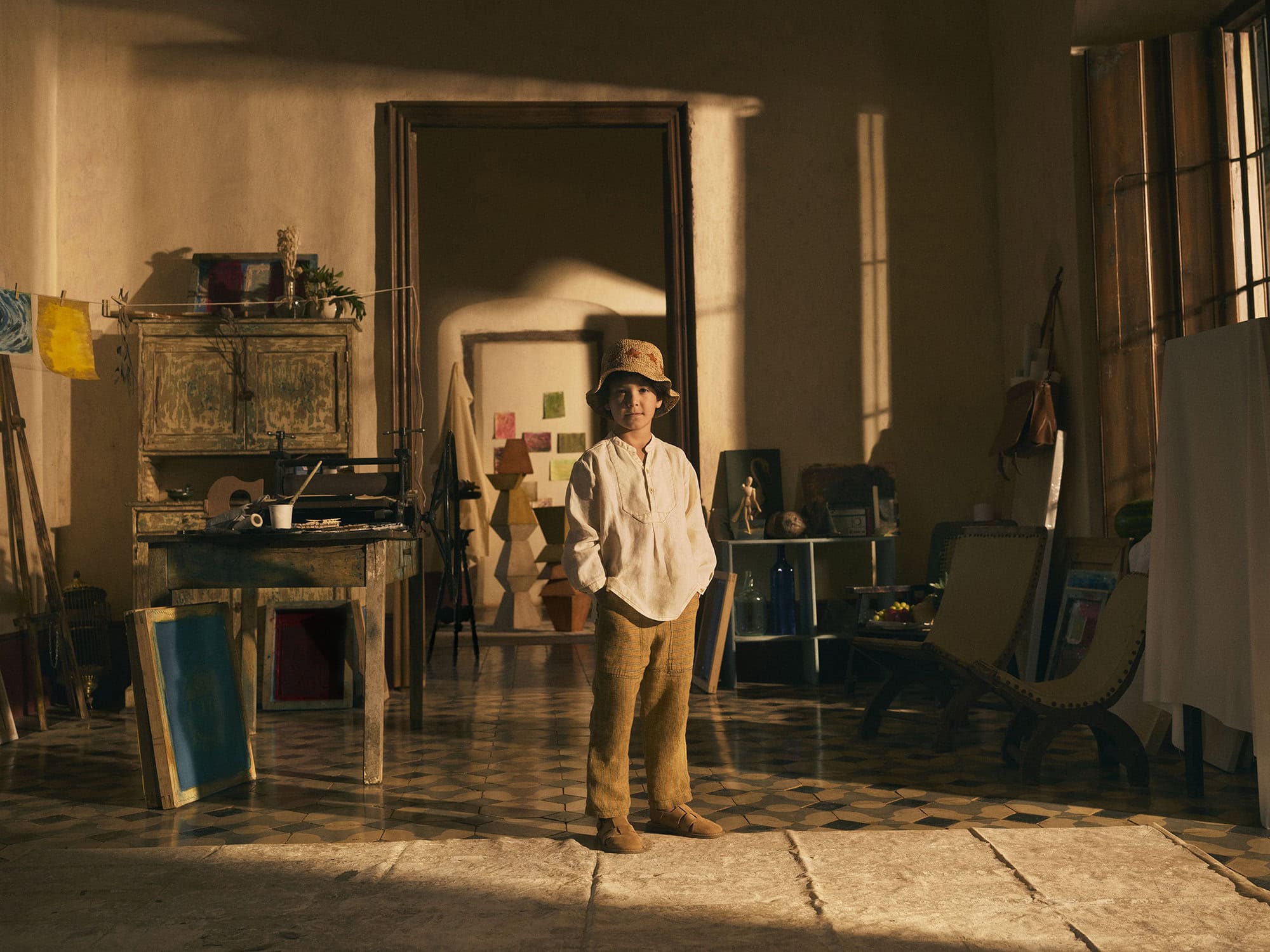 Young boy stands in an artist's studio filled with art supplies, easels, and paintings in warm, natural light, creating a vintage aesthetic.