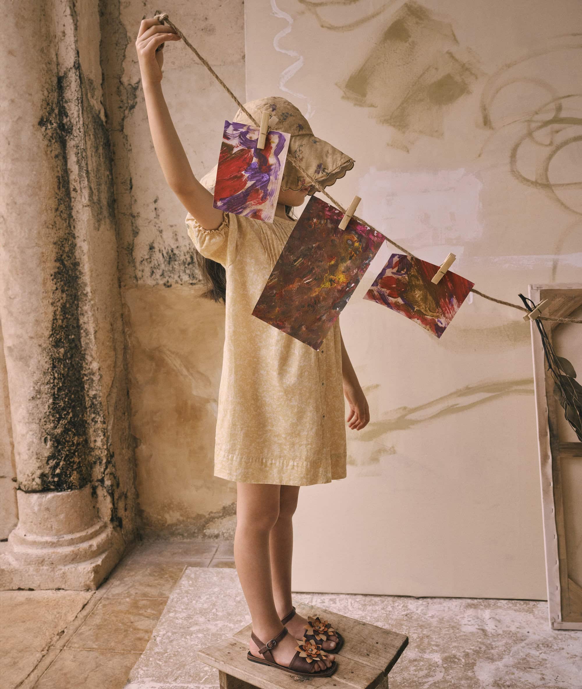 A young girl stands on a wooden stool, hanging colorful abstract paintings with clothespins on a string in a rustic indoor setting.