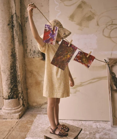 A young girl stands on a wooden stool, hanging colorful abstract paintings with clothespins on a string in a rustic indoor setting.