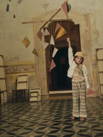 Young girl in patterned pants and hat, reaching for a mobile in a vintage interior, bathed in soft, natural light, creating a whimsical scene.