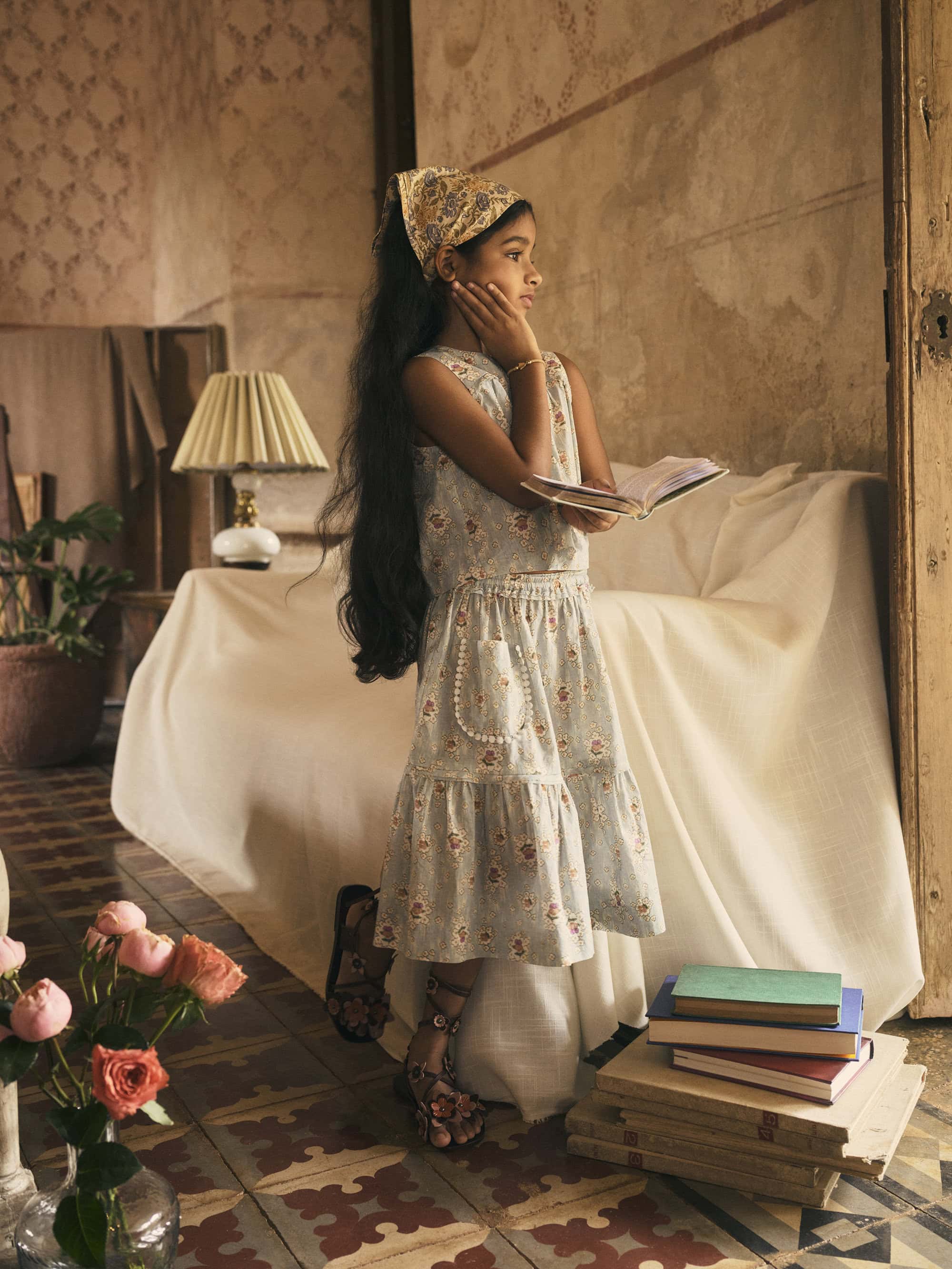 Child in a floral outfit, styled with a matching headscarf, reading a book in a vintage-style room with soft lighting and pastel colors.