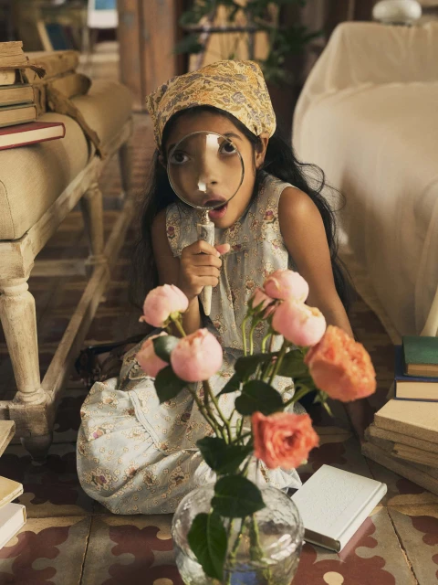 Young girl with magnifying glass sitting on a floor with a vase of pink roses and stack of books in a well-lit indoor setting.