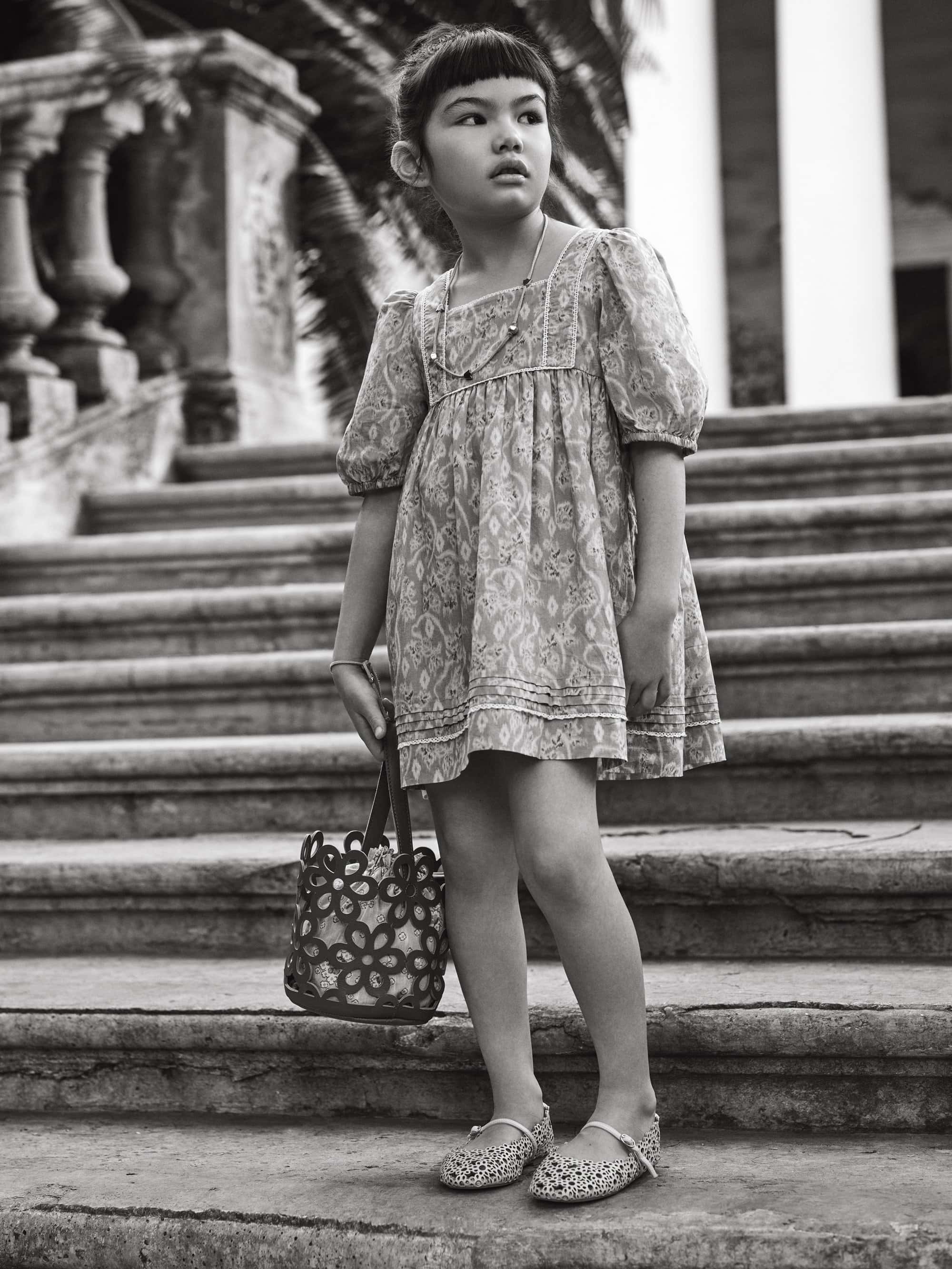 Young girl in a patterned dress stands on stone steps with a flower-cutout purse and shoes, captured in a monochrome photograph.