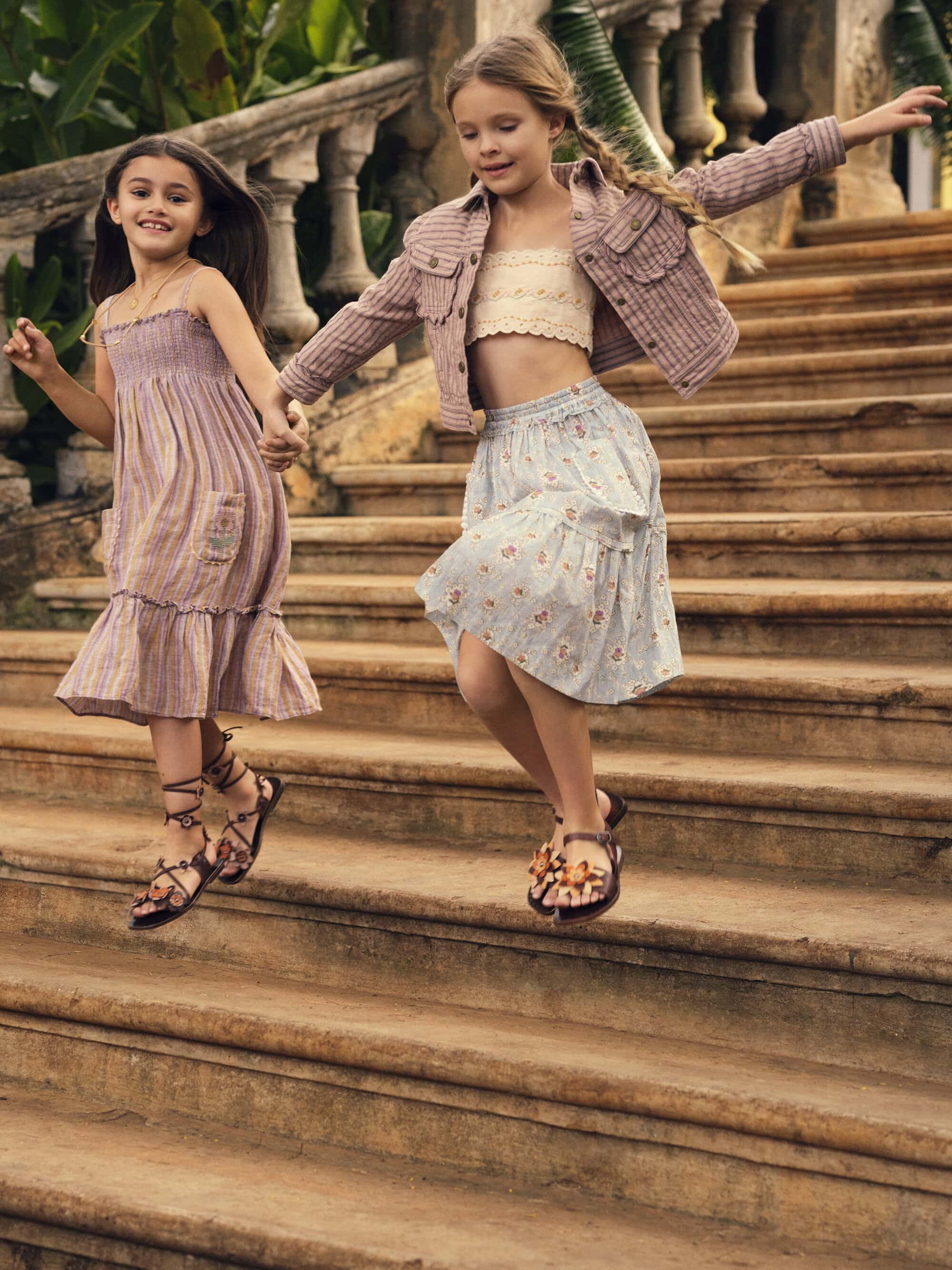 Two young girls with long hair and sandals are holding hands while descending stairs outdoors surrounded by lush green foliage.