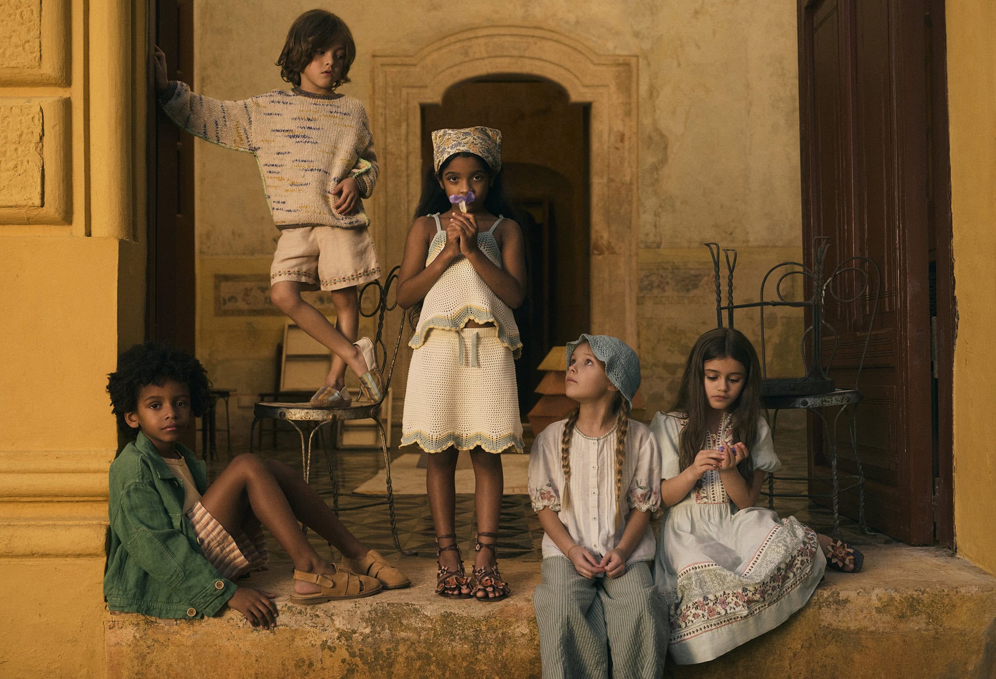 Group of five children in muted tones clothing sitting outdoors in a building entrance with tan walls and detailed architecture.