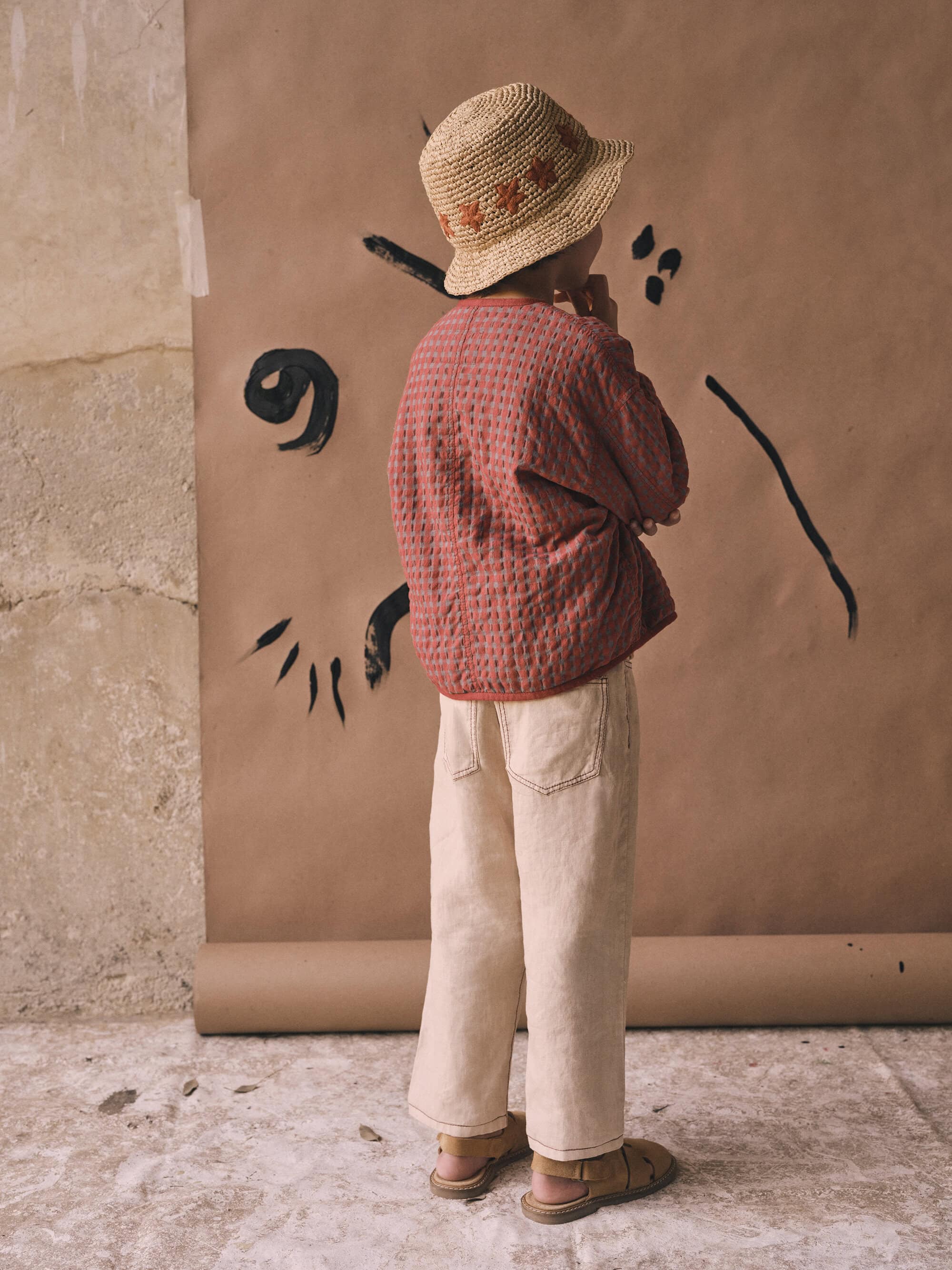 Child model in quilted jacket and wide-leg jeans poses with hand to face near a paper backdrop with abstract black ink design.