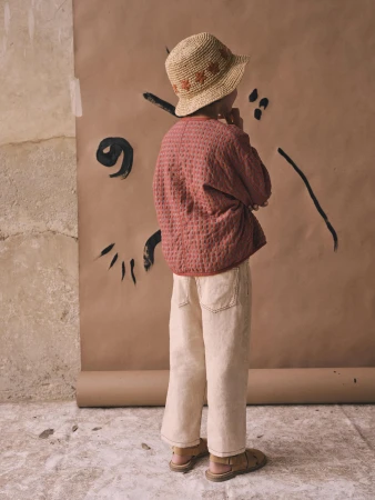 Child model in quilted jacket and wide-leg jeans poses with hand to face near a paper backdrop with abstract black ink design.