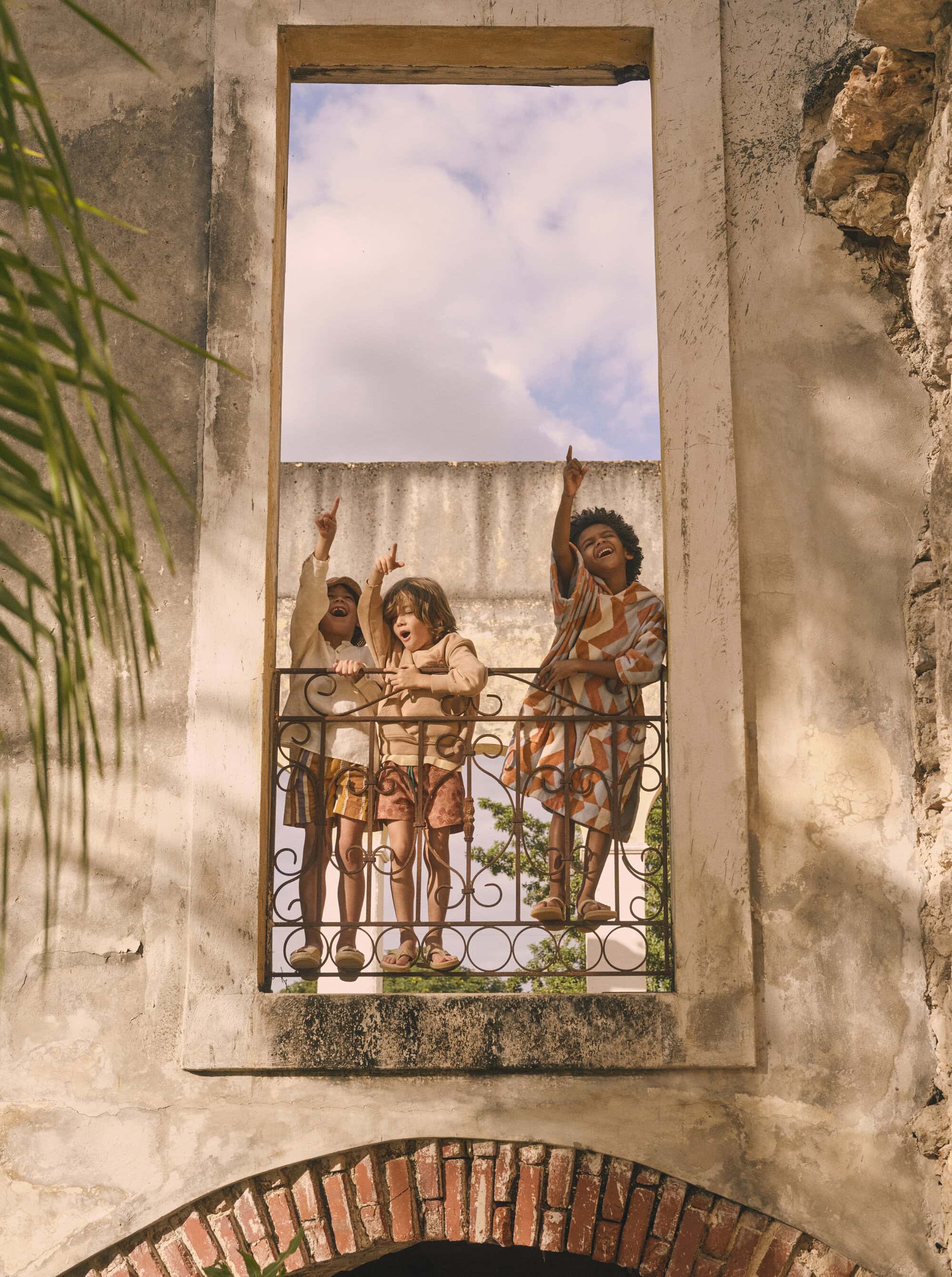 Three kids of varying ethnicities stand behind a wrought iron railing, looking up and pointing, framed by a weathered stone archway.