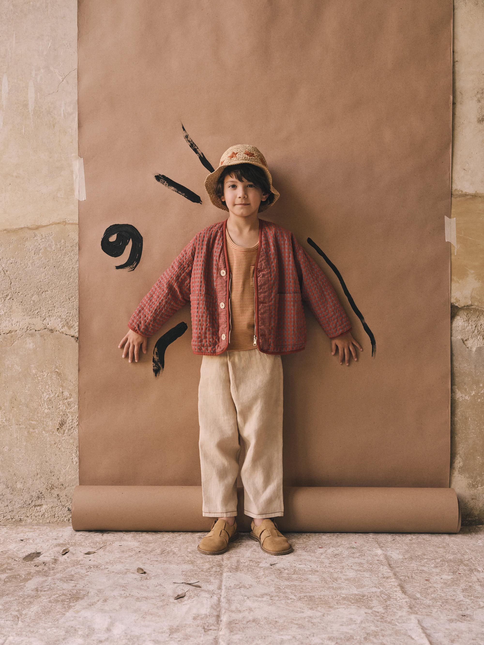 A child stands in a photo studio wearing a salmon-colored quilted jacket, tan shirt and pants, and bucket hat against a brown paper backdrop.