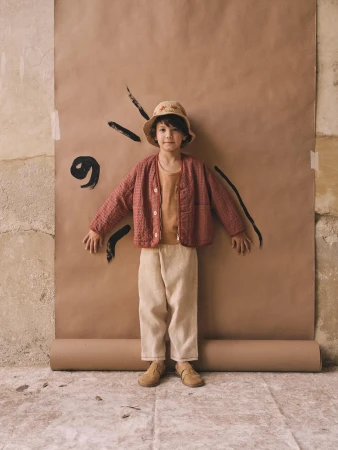 A child stands in a photo studio wearing a salmon-colored quilted jacket, tan shirt and pants, and bucket hat against a brown paper backdrop.