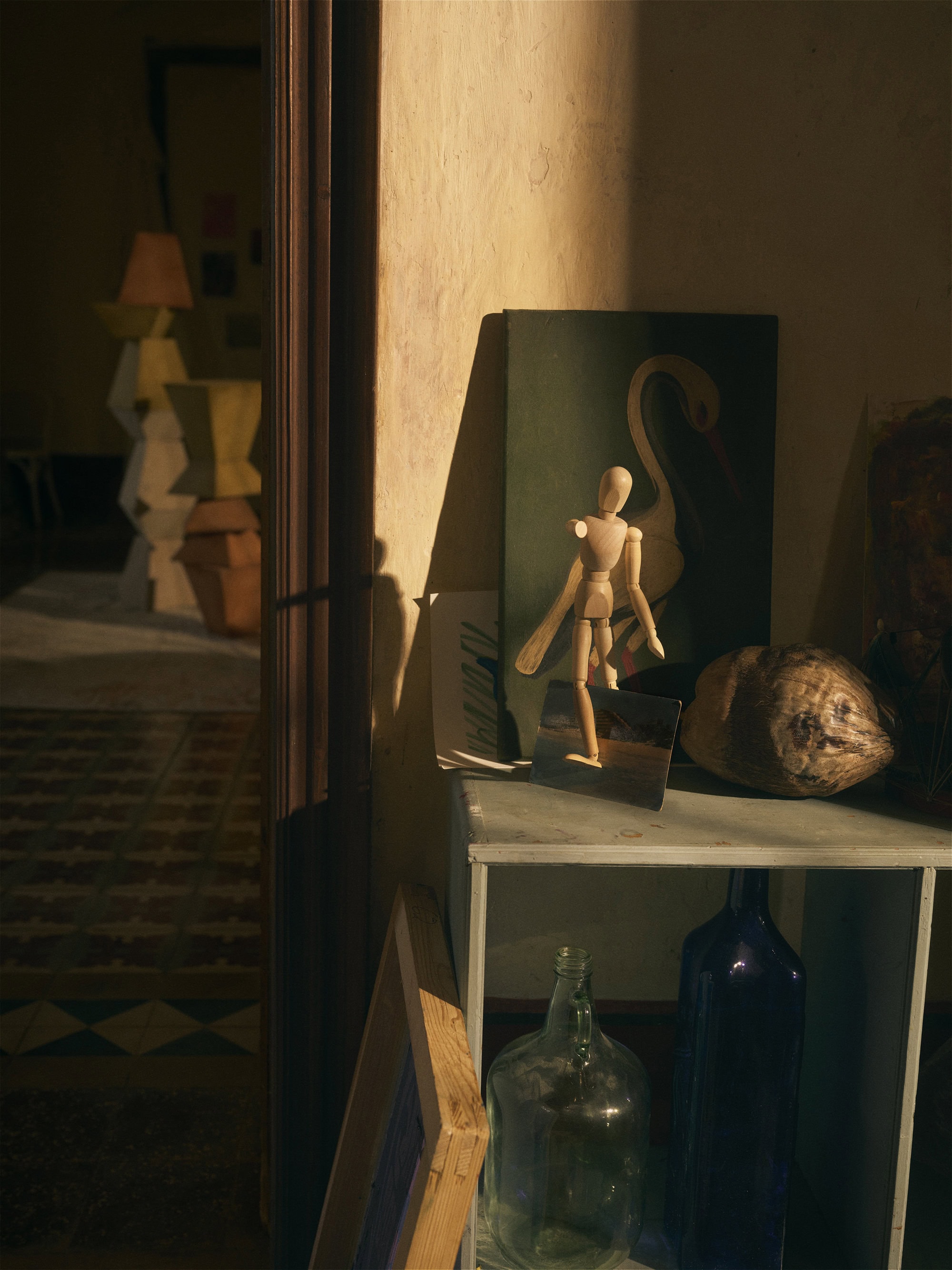 Still life featuring a wooden mannequin, paintings, and bottles on a shelf, lit by natural light in an indoor setting.
