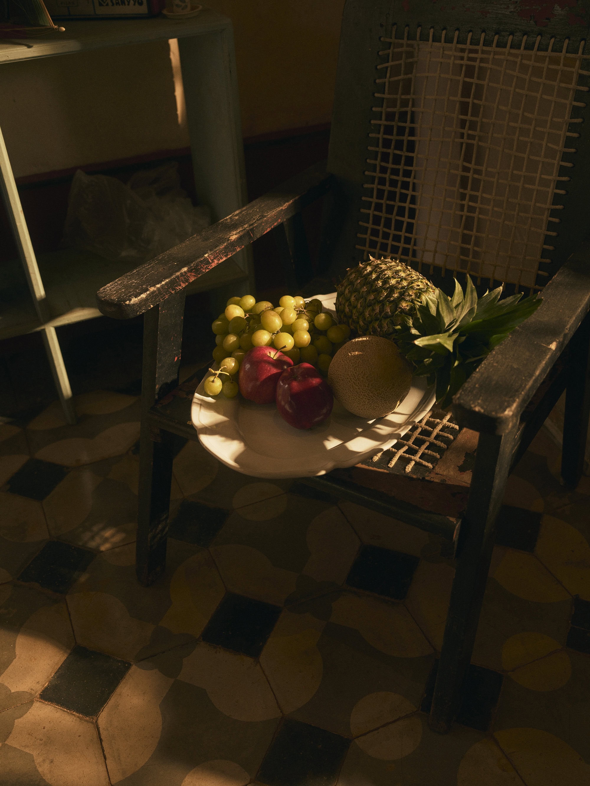Still life of pineapple, grapes, melon, and apples on a white plate, sitting on a chair with a tiled floor in the background.