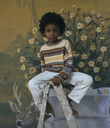 Portrait of a young boy with curly hair sitting on a weathered ladder, set against a hand-painted mural with floral and deer motifs.