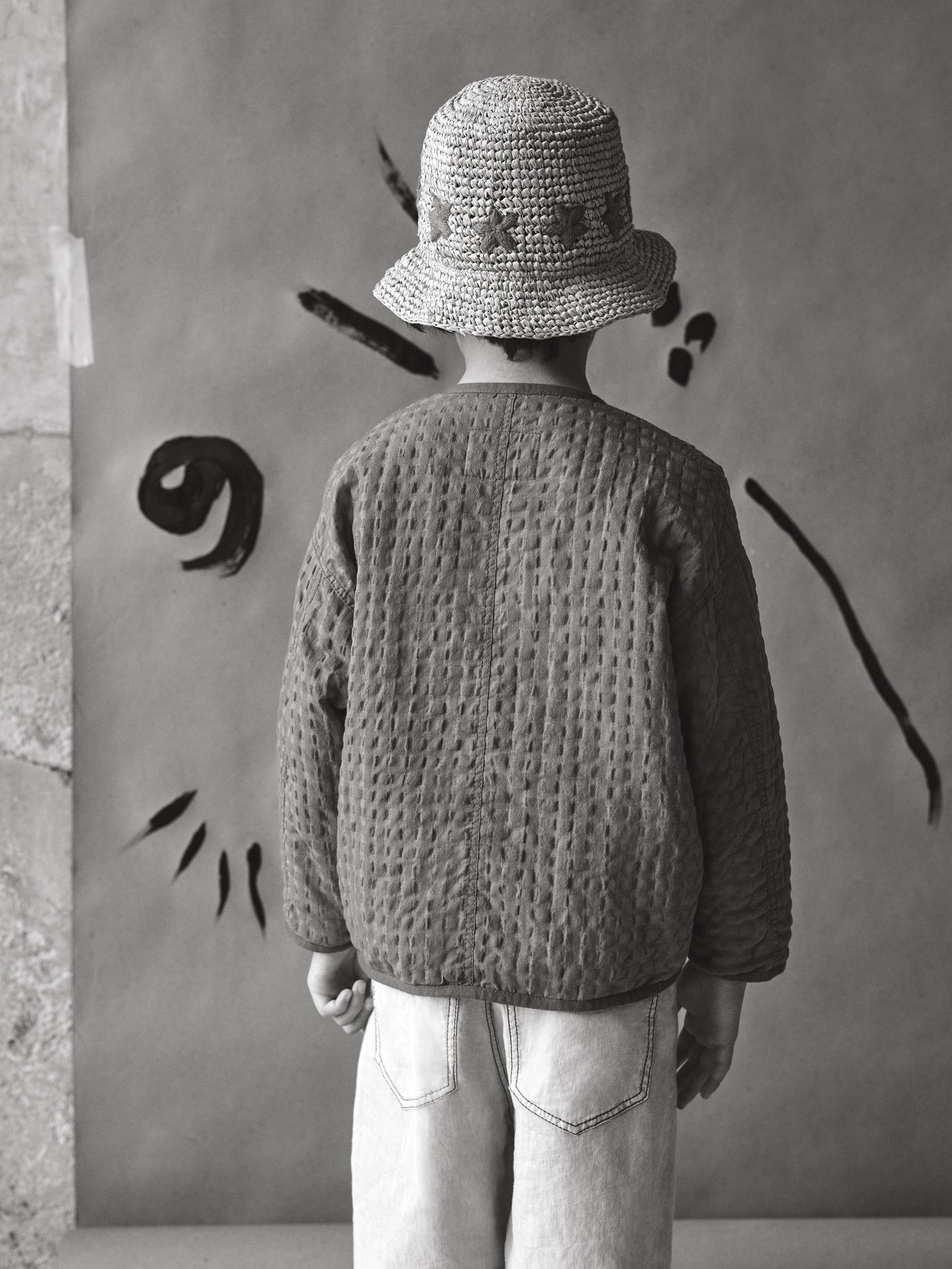 Back view of a child in a straw hat and quilted jacket against an abstract painted backdrop in black and white.