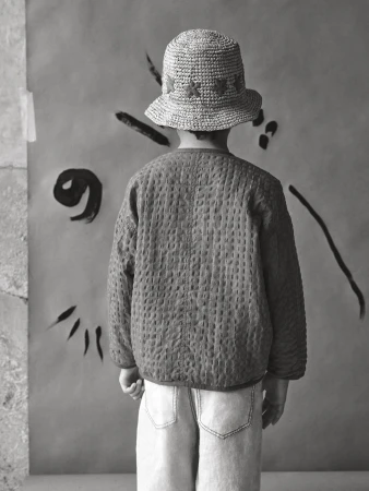 Back view of a child in a straw hat and quilted jacket against an abstract painted backdrop in black and white.