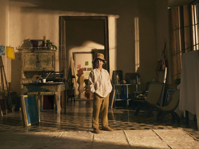 A young child wearing a bucket hat, light shirt, and trousers standing in a sunlit artist studio.