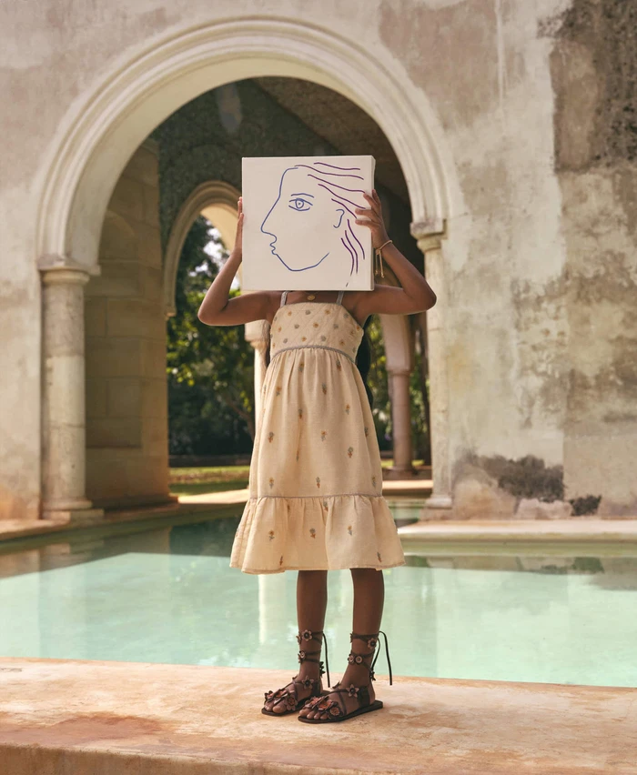 Child wearing a cream embroidered dress holding a canvas artwork in front of their face by a pool and arches.