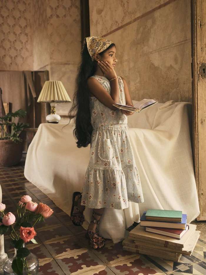 Young girl holding a book wearing a floral top and skirt with a headscarf in an interior room with a lamp and stacked books.