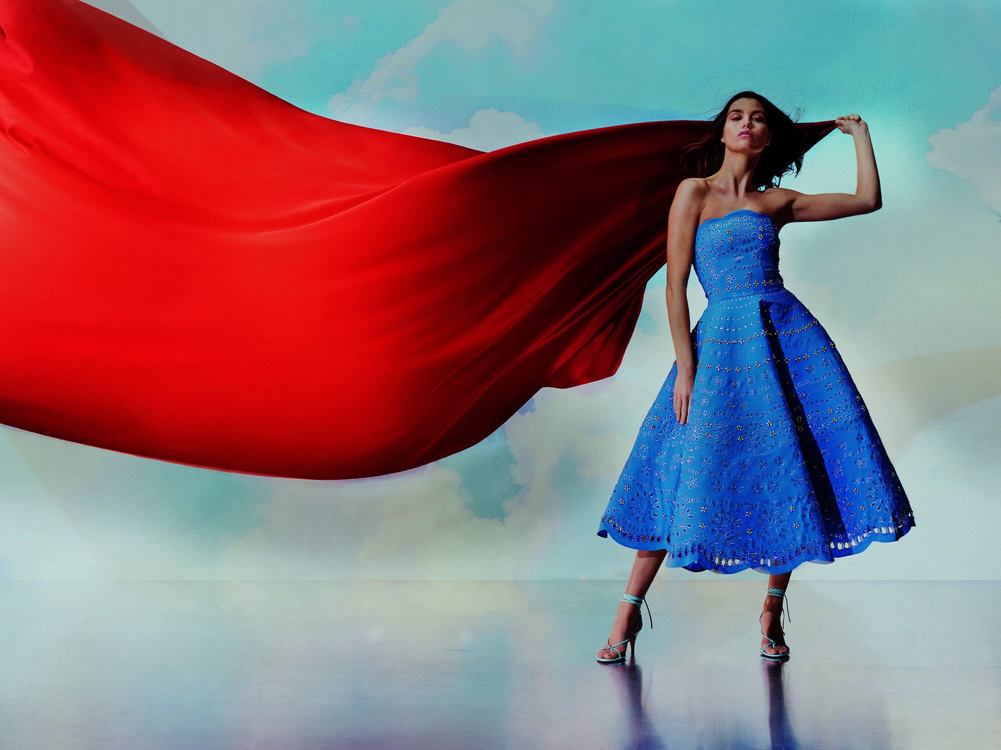 A woman in a blue dress poses in front of a sky backdrop, with a large red fabric blowing around her, wearing heels.