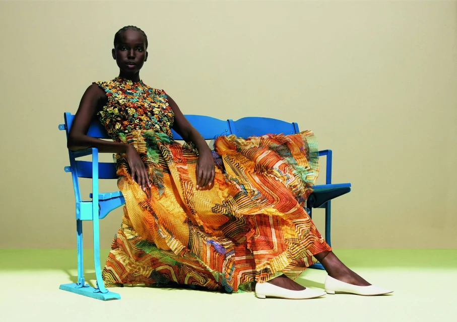 A fashion shot of a Black woman in a flowing yellow, orange, green dress sitting on a blue metal bench against a light yellow background.
