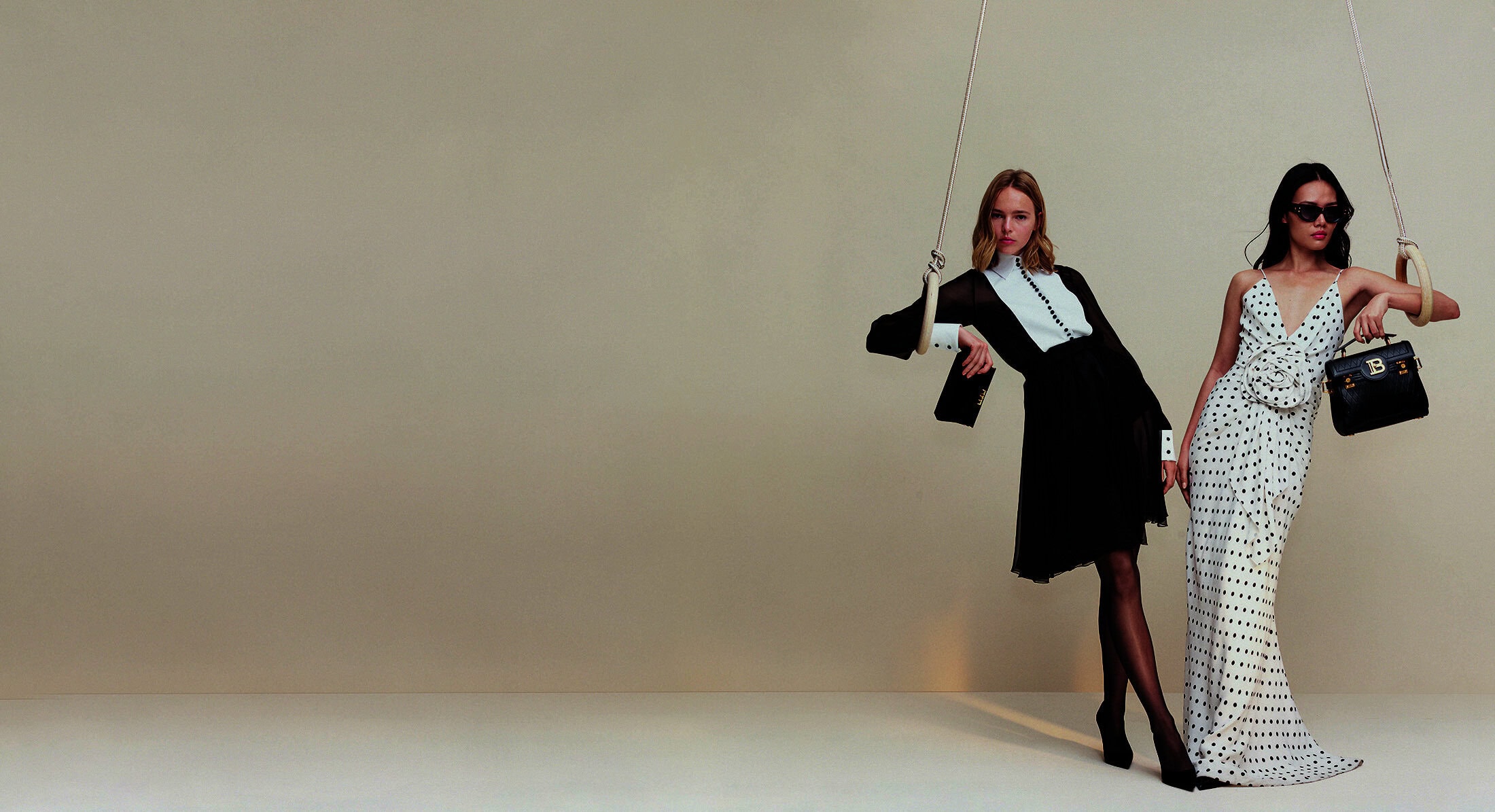 Two women in dresses, one in black and one in white with black polka dots, are hanging from metal rings in a minimalist setting.