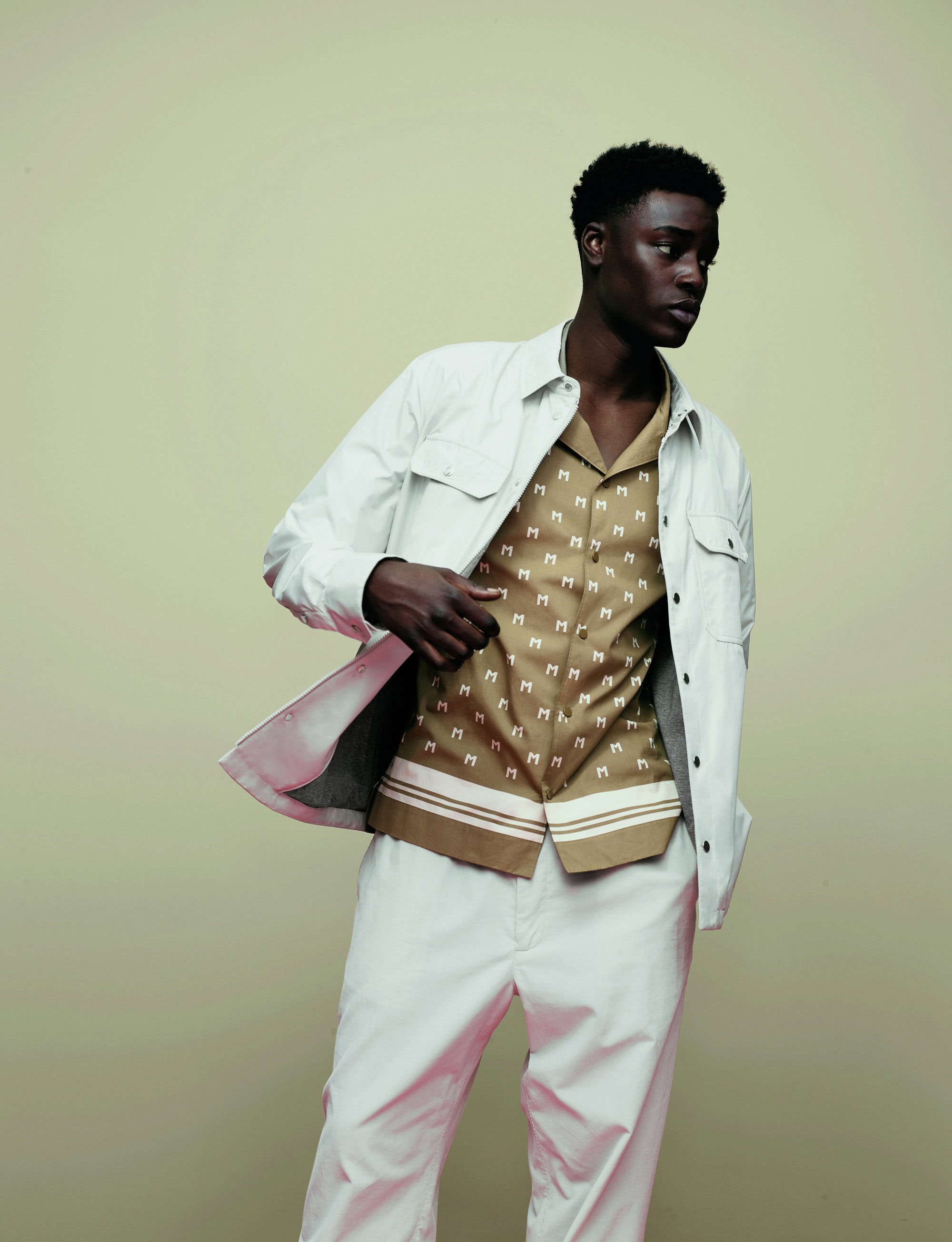 Dark-skinned male model poses indoors wearing an open white overshirt, patterned button-down shirt, and light pants against a solid backdrop.