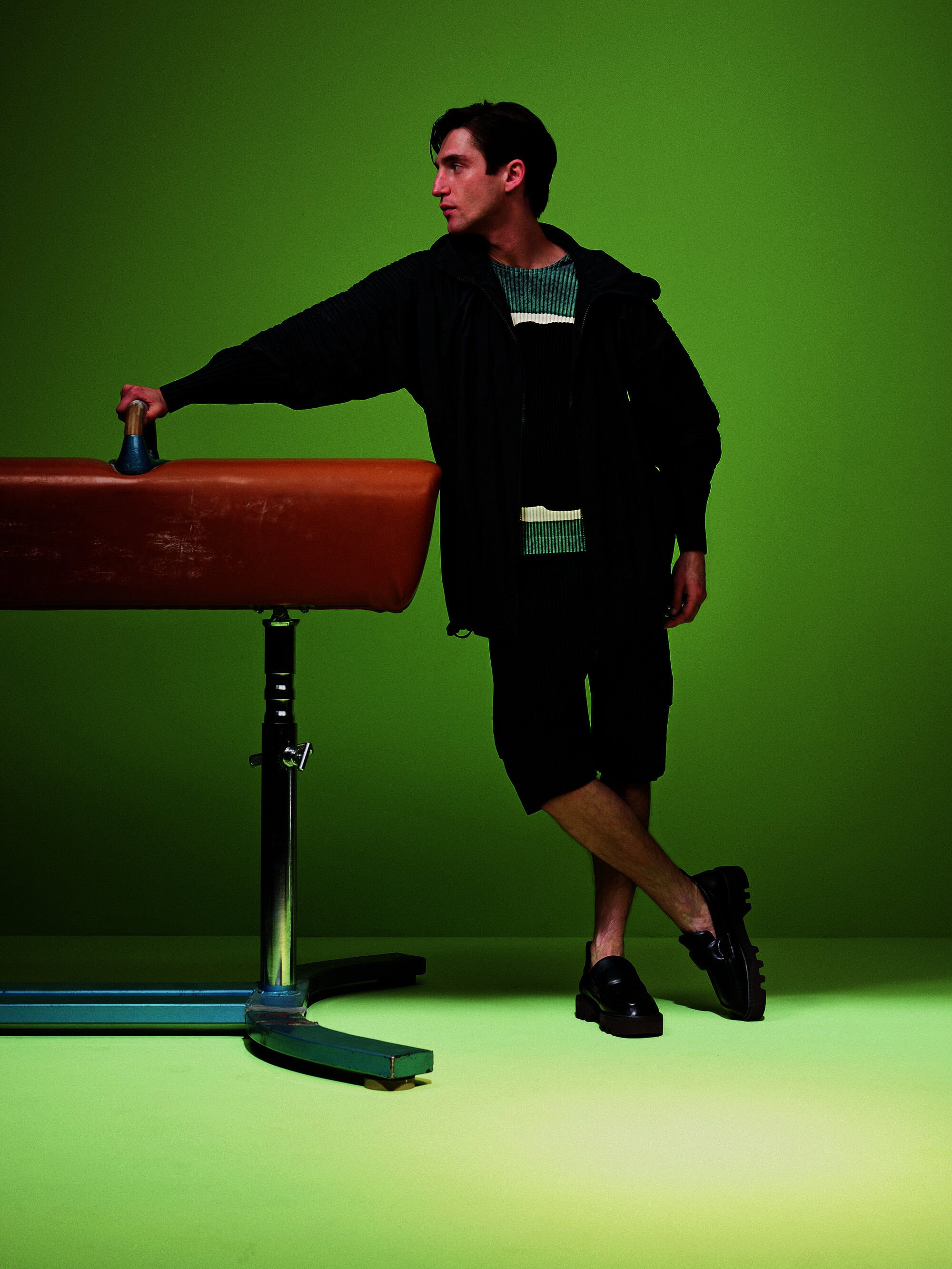 A young adult male leans against a brown leather gymnastics vault horse in a bright photo studio with a green background and floor.