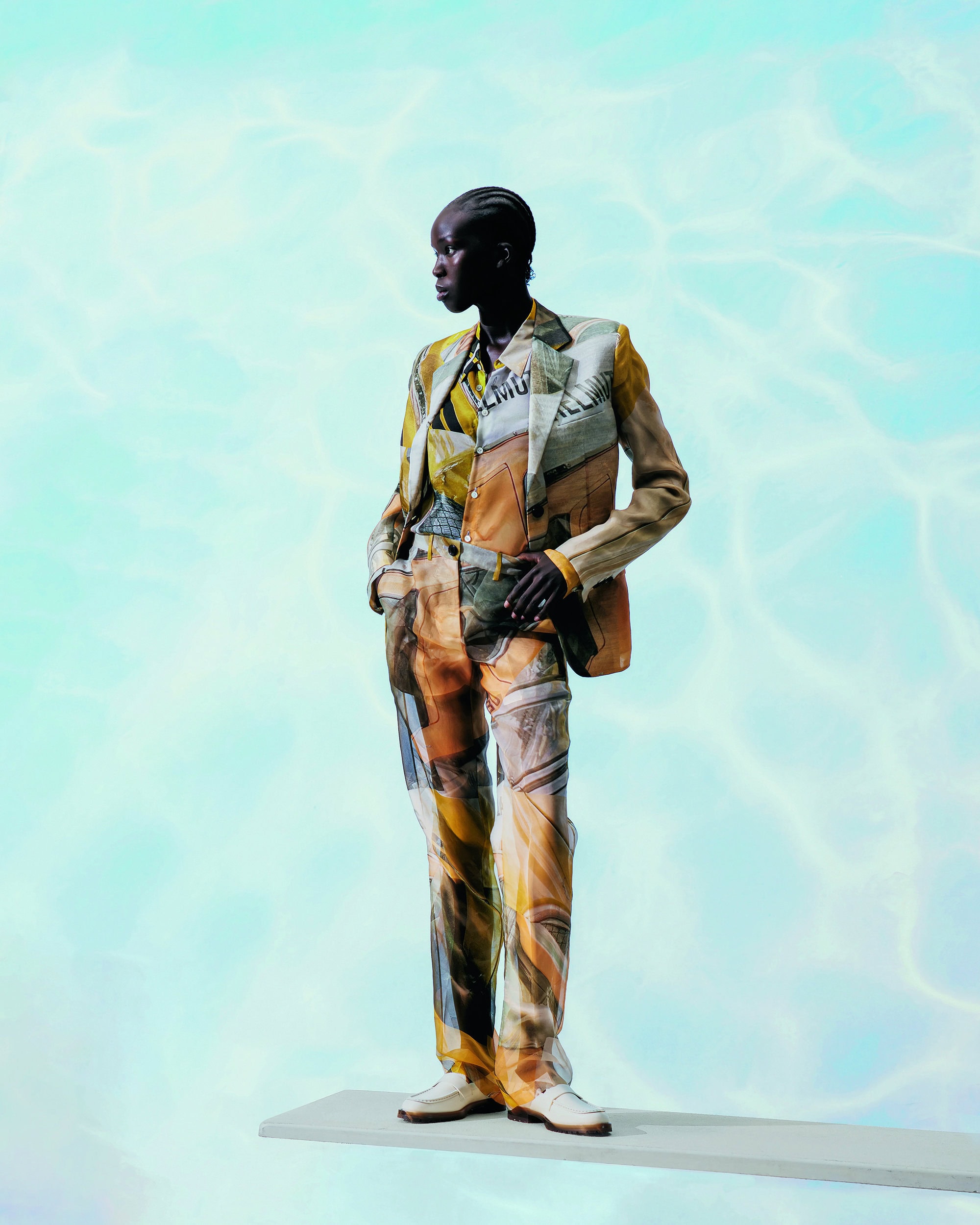 An African male fashion model stands in a printed suit with a blue water-patterned background, showcasing modern, stylish apparel in a studio setting.
