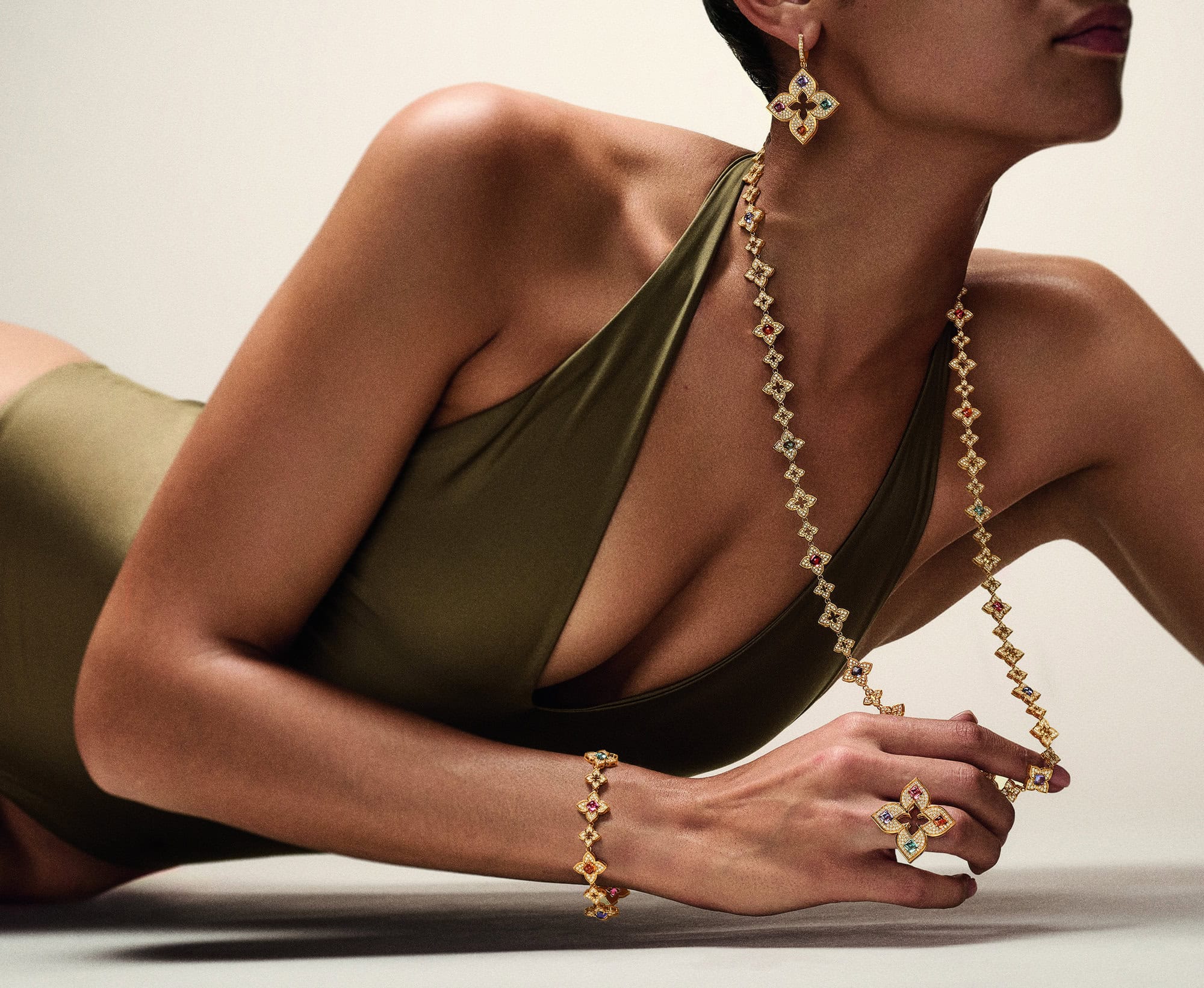Woman in an olive swimsuit laying down wearing an earrings, necklace, ring, and bracelet with a flower design on each jewelry piece.