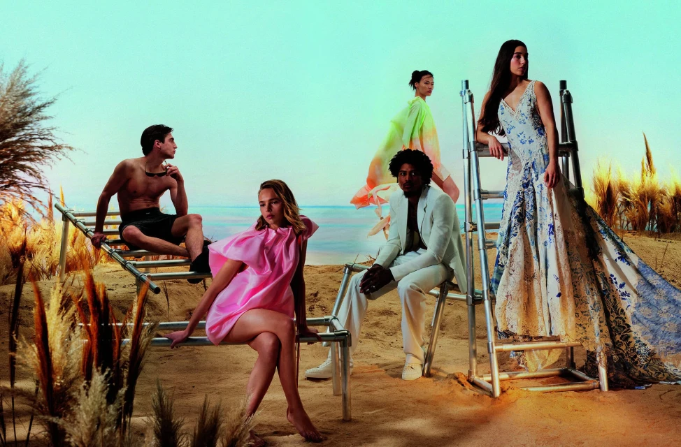 Fashion editorial shot of five people posing on a beach, wearing a mix of swimwear, dresses, and suits in a light and airy aesthetic.