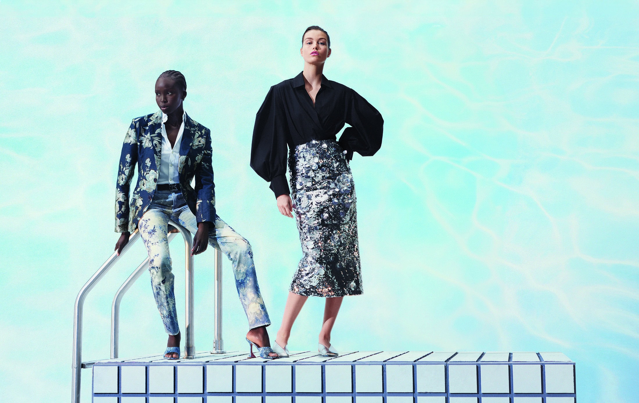 Two women pose on a tiled platform in designer clothing, one seated in a floral suit, and the other standing in a black top and silver skirt.