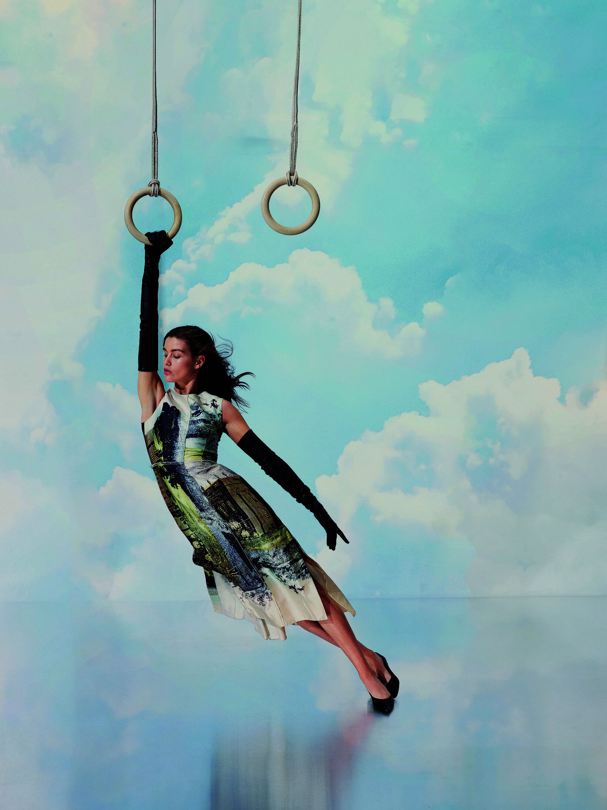 Woman in dress and gloves hanging from gymnastic rings against a cloudy background. Fashion, acrobatics, and surreal composition.