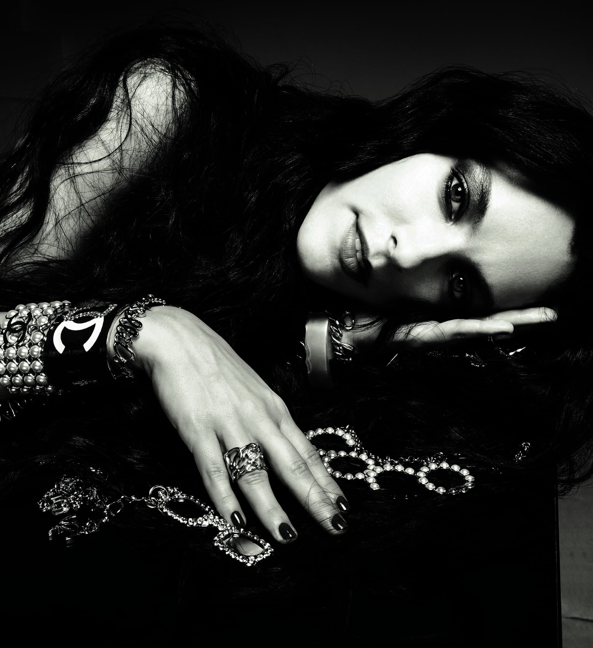 Close-up portrait of a woman with dark wavy hair wearing layered Chanel pearl and logo bracelets, resting her head on her hand.