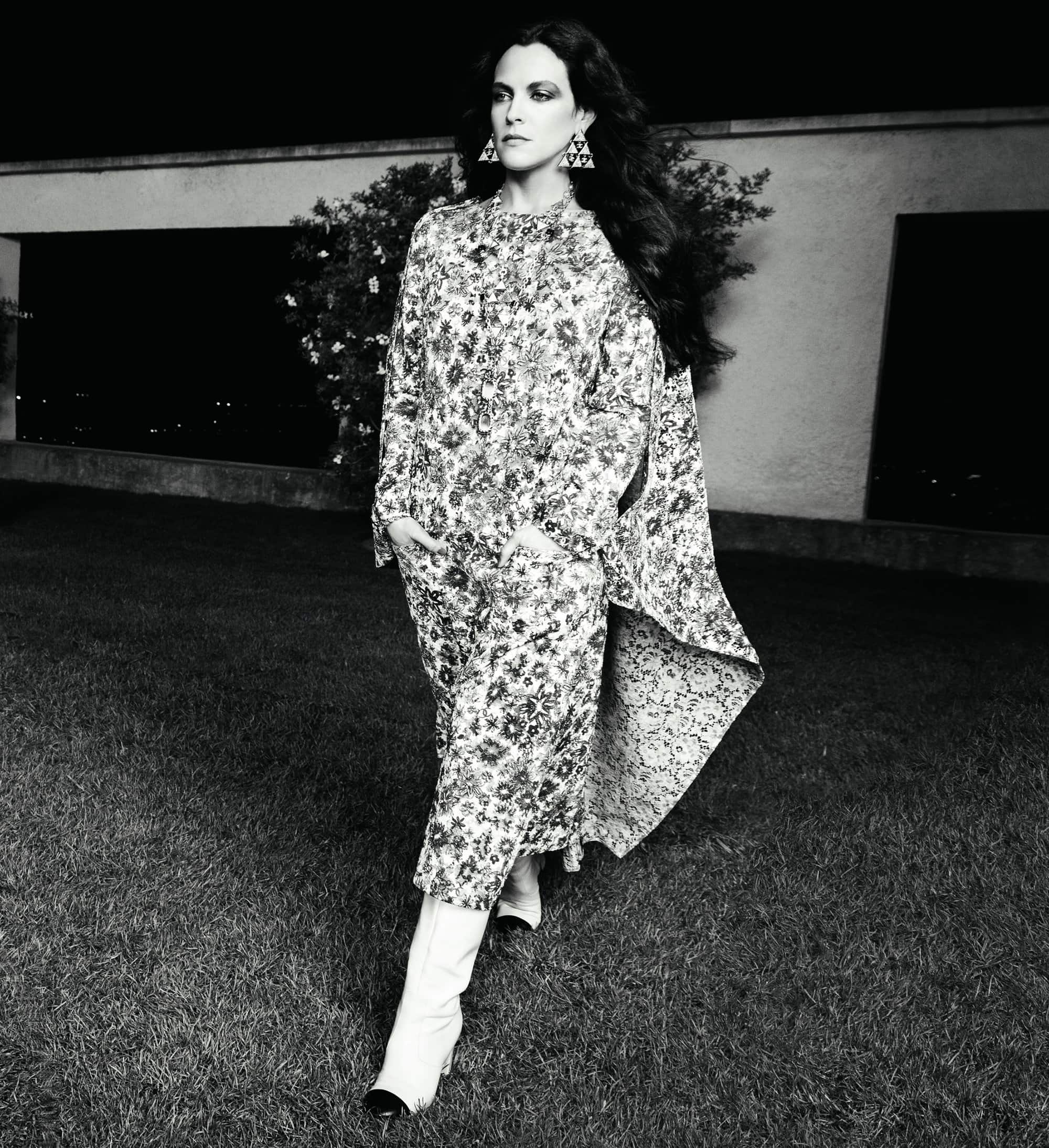 Monochrome portrait of a stylish woman in a floral-print dress and white boots, posing on a lawn in front of a building at night.