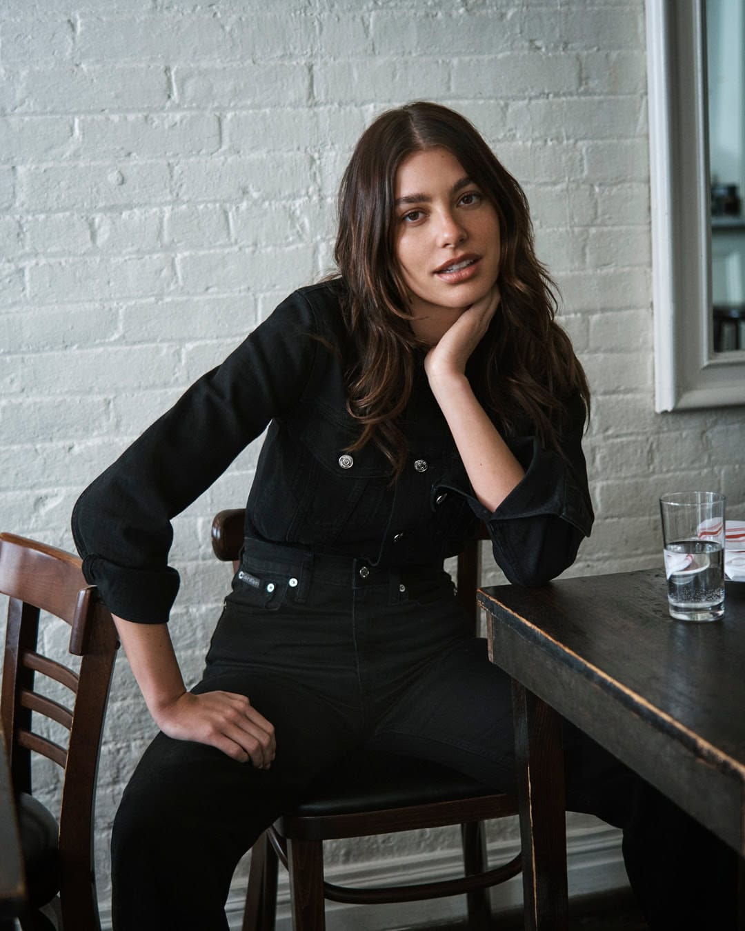 Brunette woman in a black denim jacket and jeans sitting at a table with hand to face and a brick wall background.