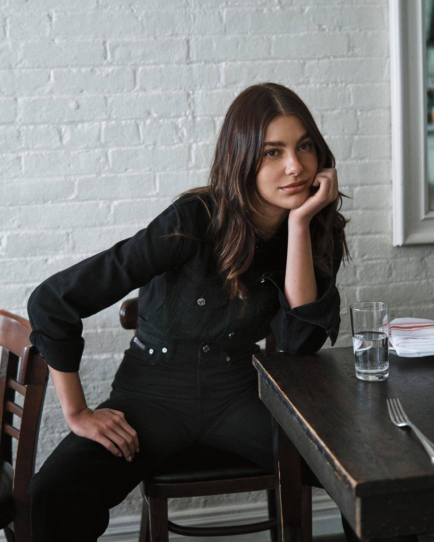 Young woman in black denim sits at a table in a restaurant with a glass of water and a fork, looking directly at the camera.