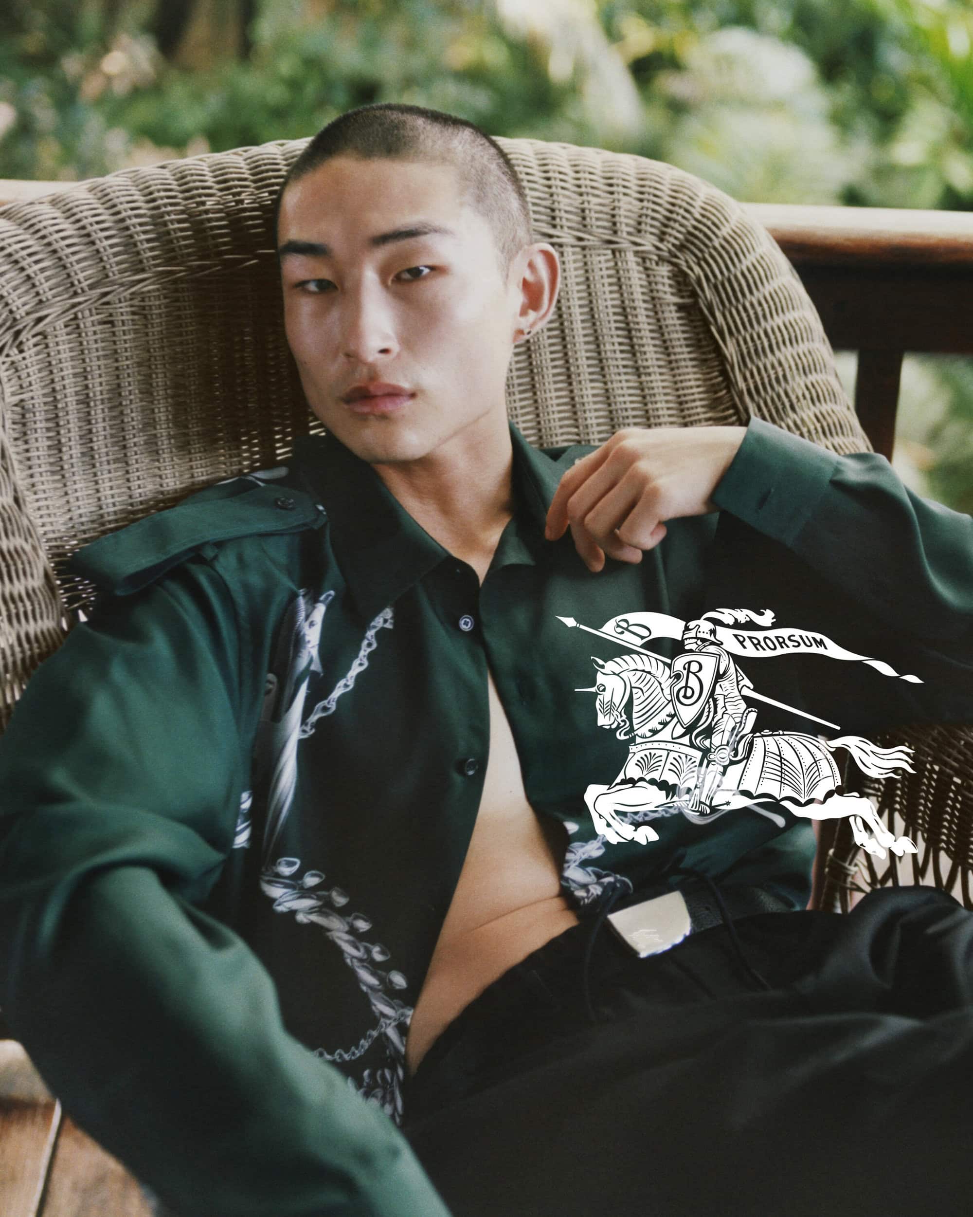 Asian male model with shaved head poses in wicker chair in dark green open collared shirt and black pants with greenery background.