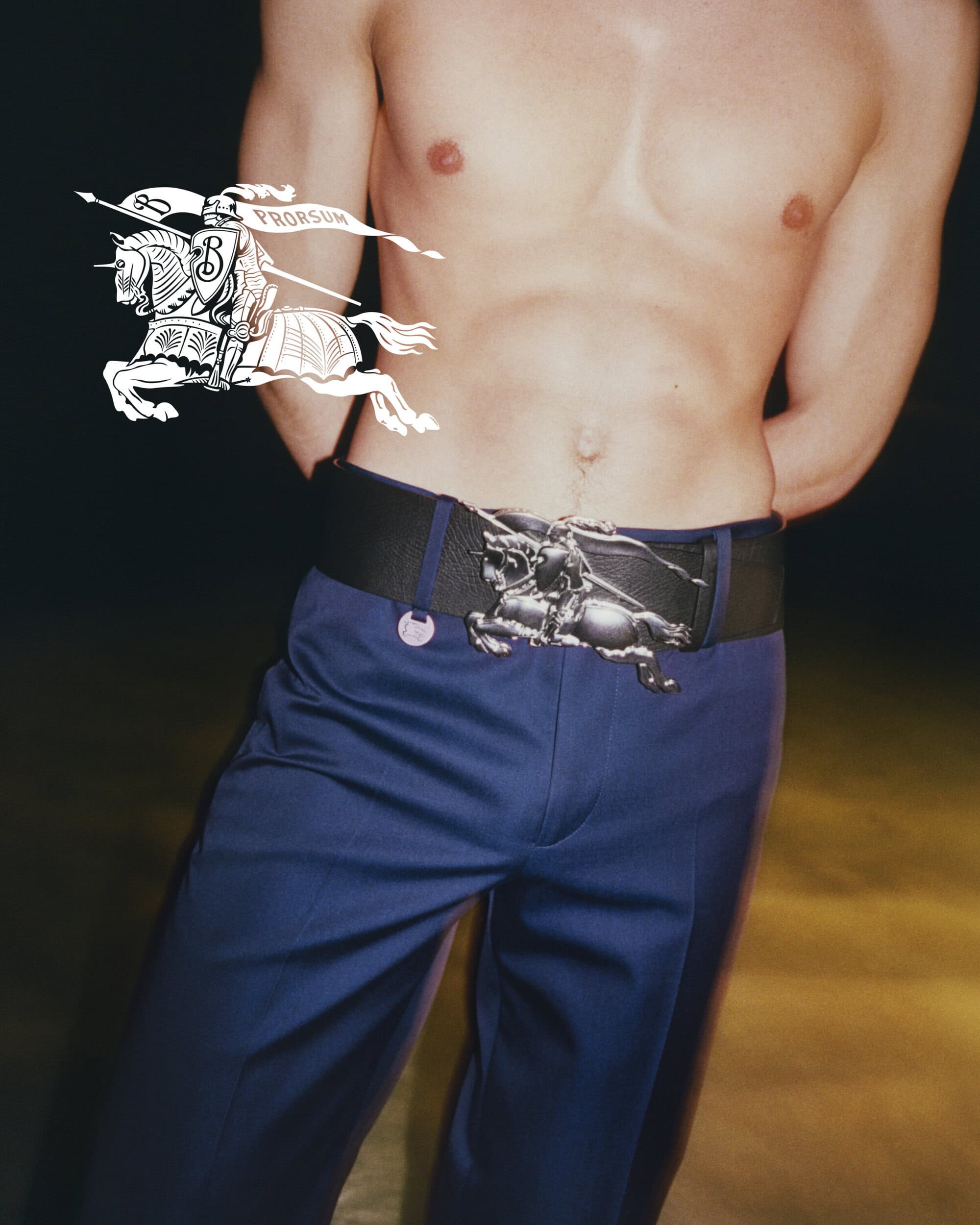 Torso of a topless male model wearing blue pants and a wide belt with a silver-toned decorative buckle in a studio shot.