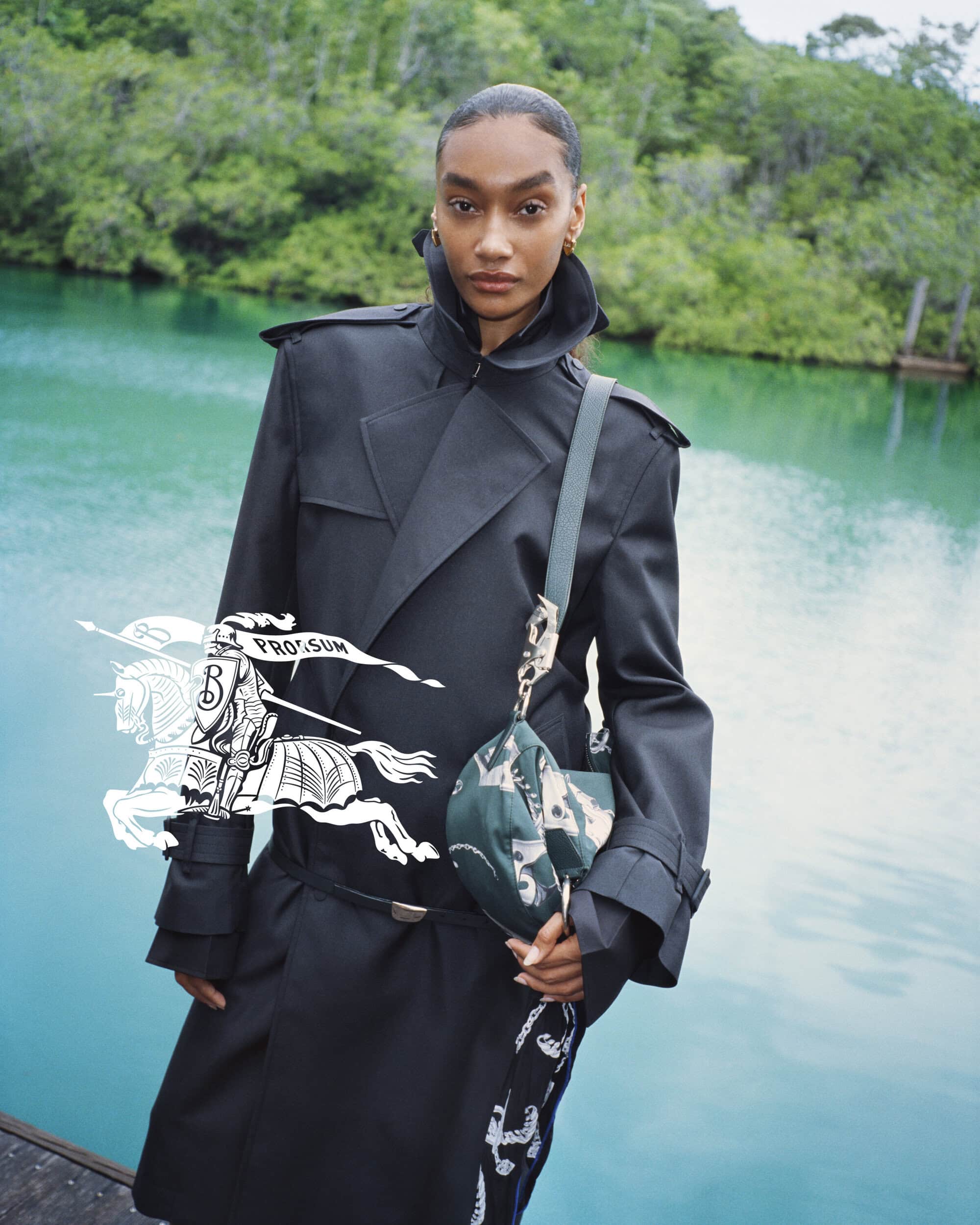 Fashion shot of a female model wearing a black trench coat with a camo handbag, posing in front of a body of water and lush greenery.
