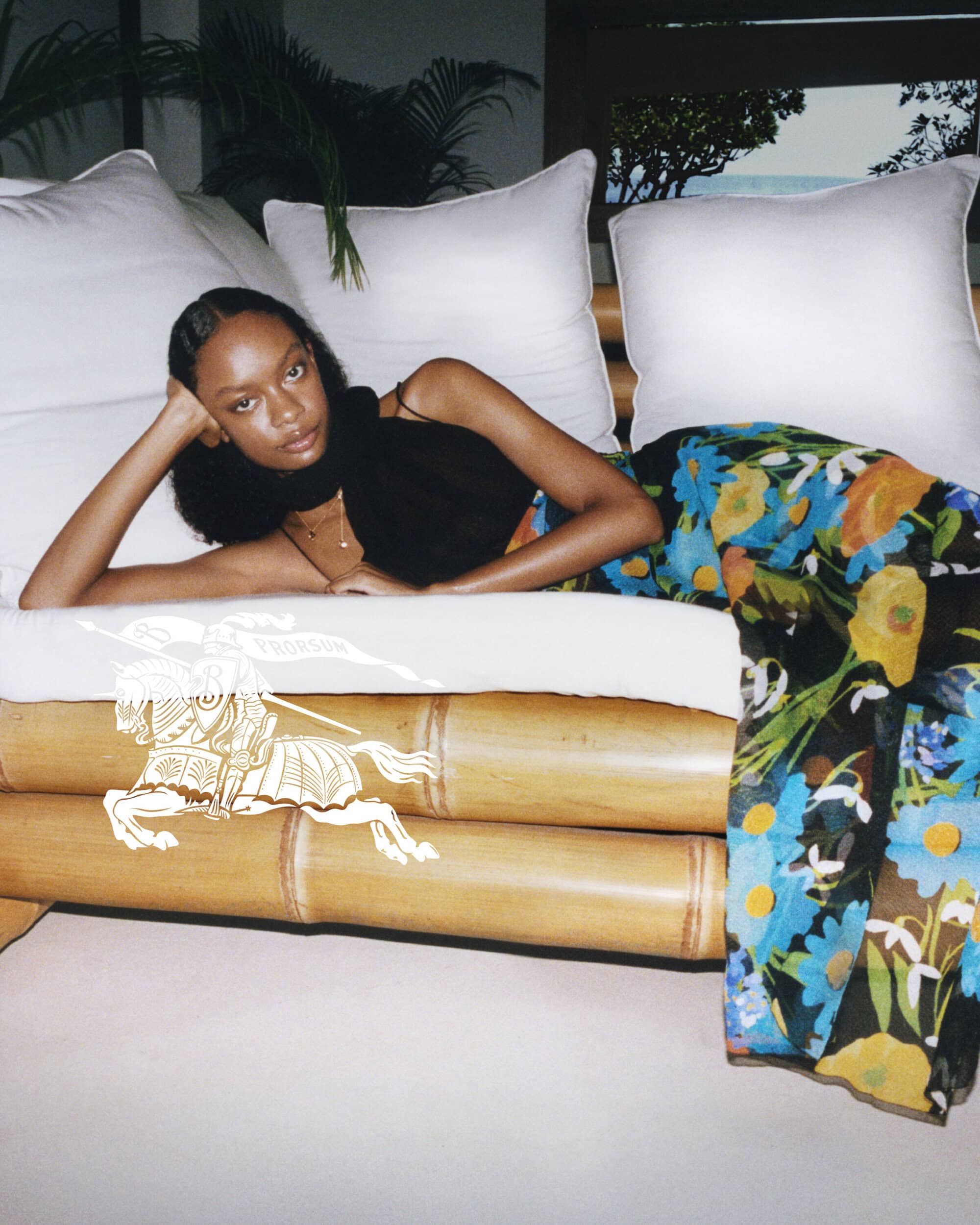 Elegant woman in floral skirt lounges on bamboo daybed by ocean view. Sophisticated indoor fashion shot with neutral color palette.