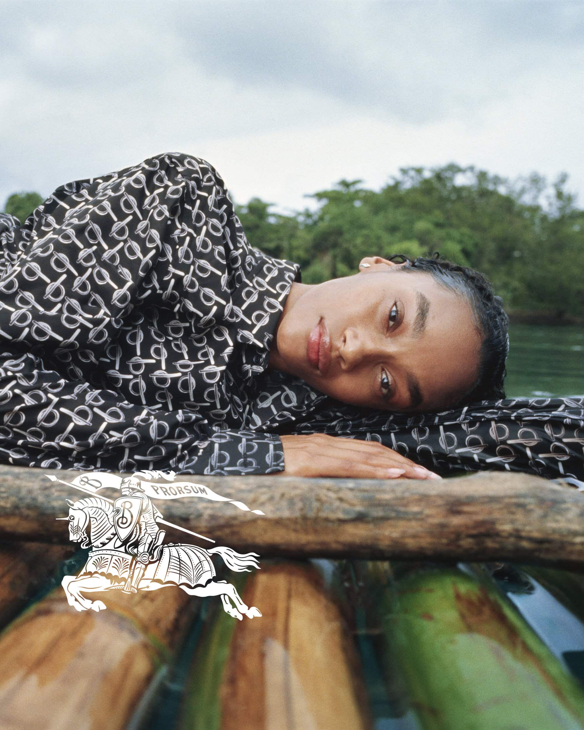 An adult model poses outdoors in a black and white patterned shirt while laying on a wooden platform with a river background.