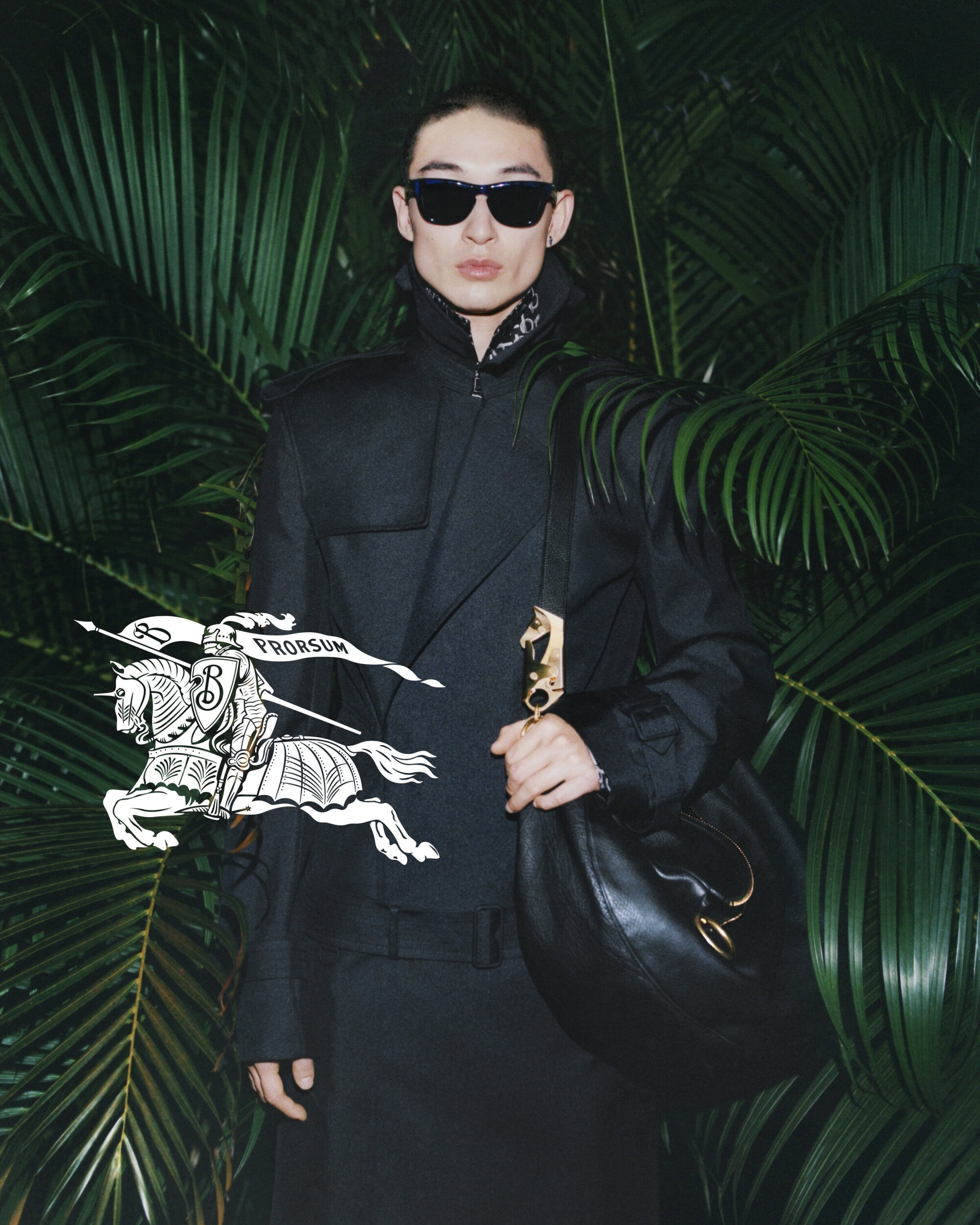 A person in a black jacket with dark sunglasses stands against a backdrop of lush green palm fronds, holding a black leather bag.